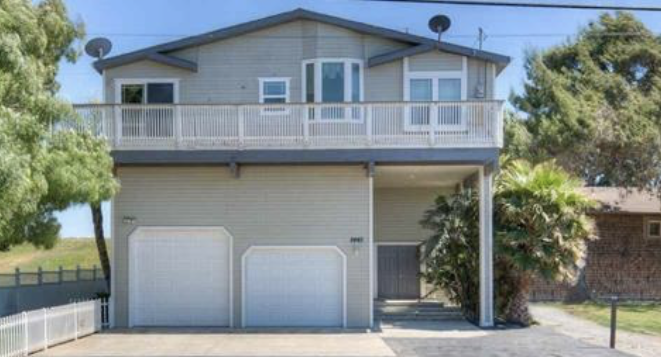 A three-story house with a garage on the ground level, a balcony with a white railing on the middle level, and satellite dishes on the roof. The house has beige siding and is surrounded by trees.