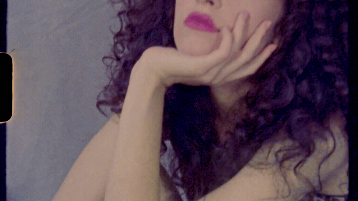 A woman with curly dark hair and pink lipstick resting her chin on her hand.