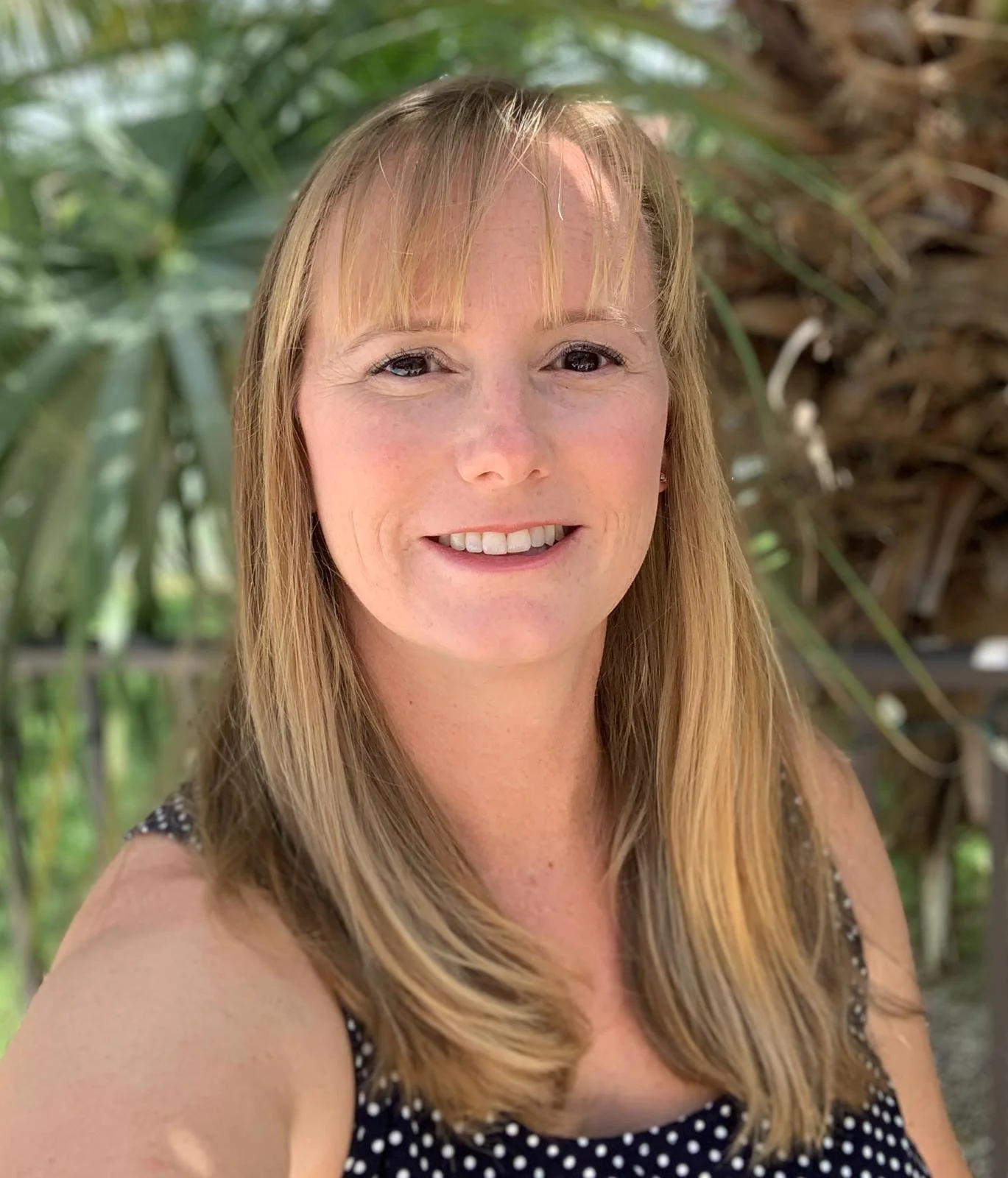 A woman with blonde hair and freckles smiling outdoors among green plants and trees.