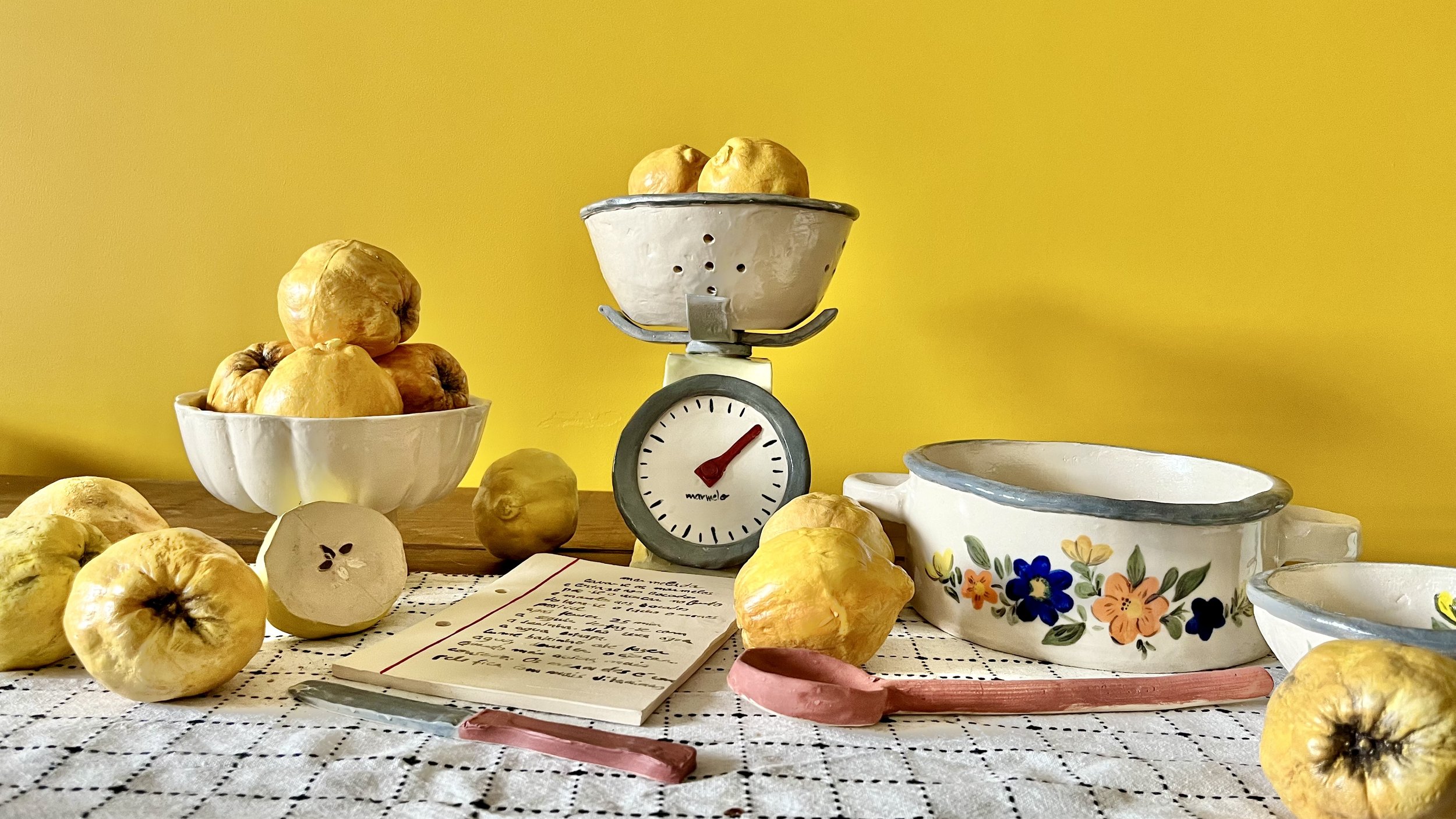 A still life scene with a yellow background displaying a kitchen scale with yellow quinces, some in a white bowl and others scattered across a checkered cloth on a wooden surface, alongside a floral ceramic bowl, a handwritten note, and a small pink ceramic spoon.