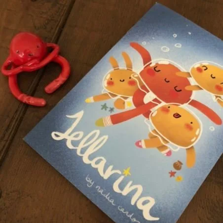 A children's book titled 'Jellarina' with cartoon jellyfish on the cover, placed on a wooden surface next to a red octopus toy.