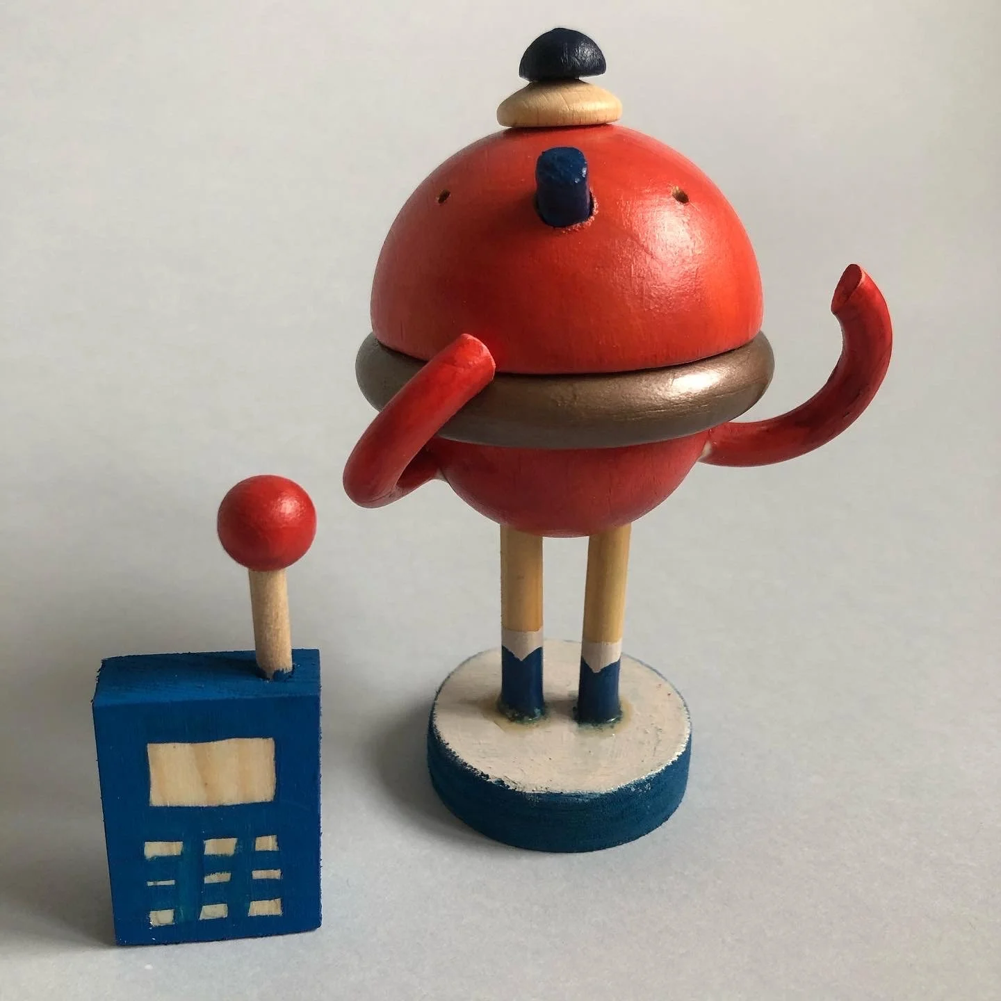 Colorful wooden robot toy with a red body, blue and white legs, and black and red accessories, standing next to a blue block with a white rectangle.