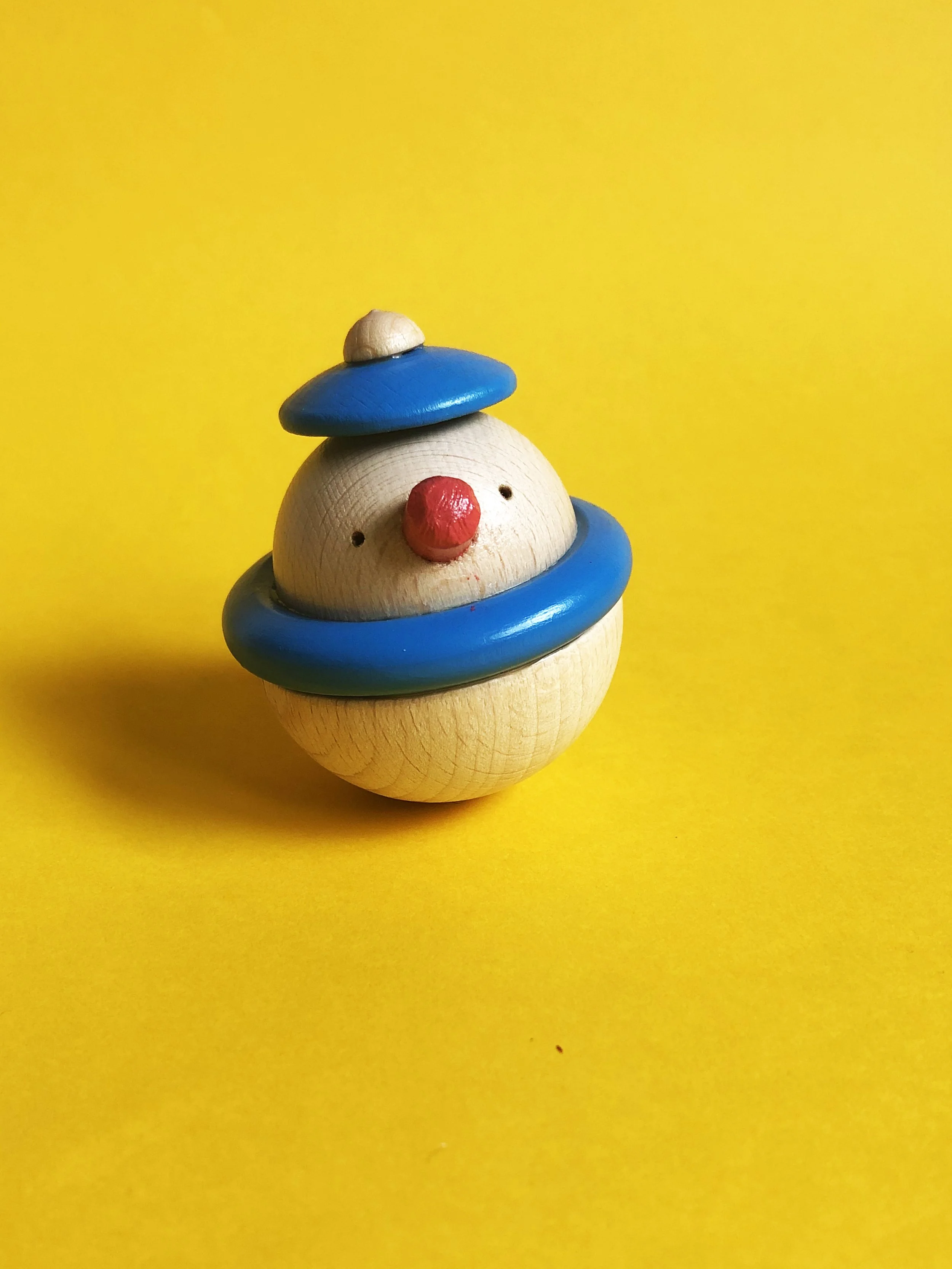 A small wooden clown figurine with a red nose, blue hat, and blue ring around its neck, against a bright yellow background.