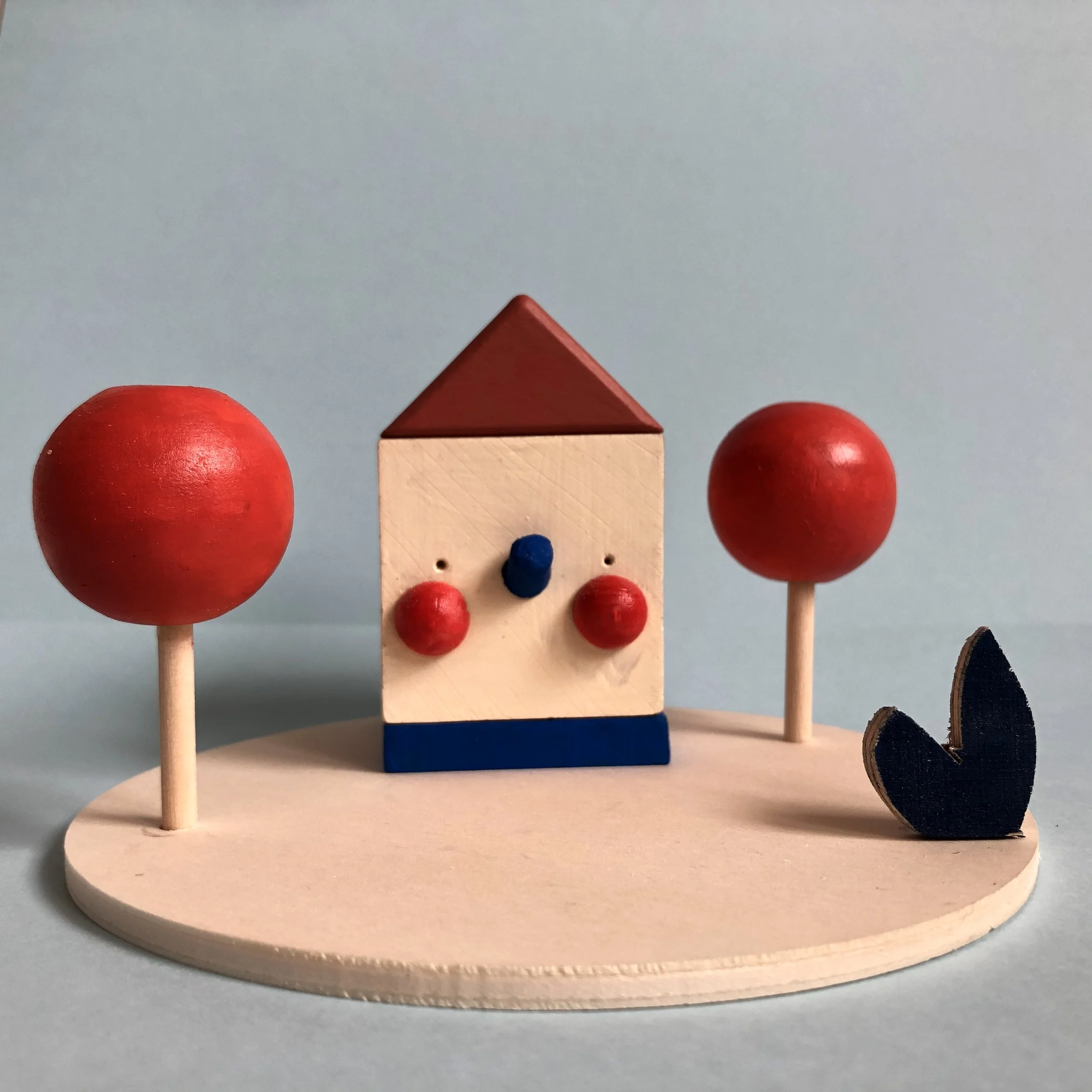 Wooden toy house with a triangular roof, two red sphere trees on either side, set on a round wooden base.