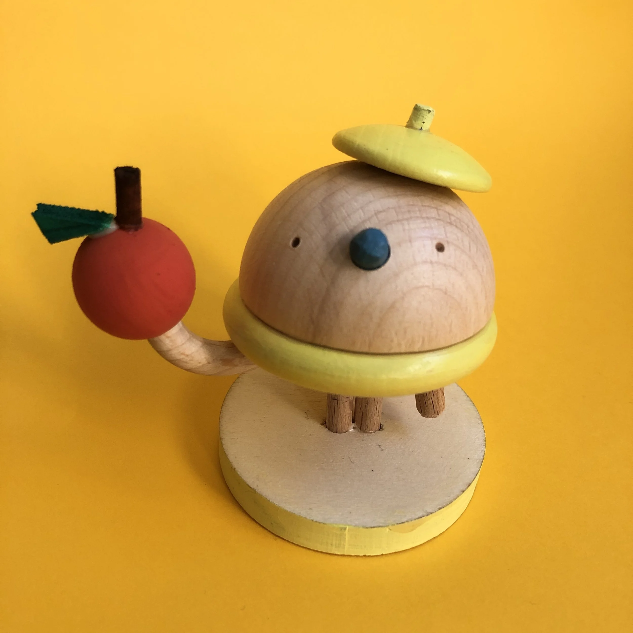 Wooden bird with a yellow beak and yellow hat next to a red apple with a green leaf and a brown stem, set against a yellow background.