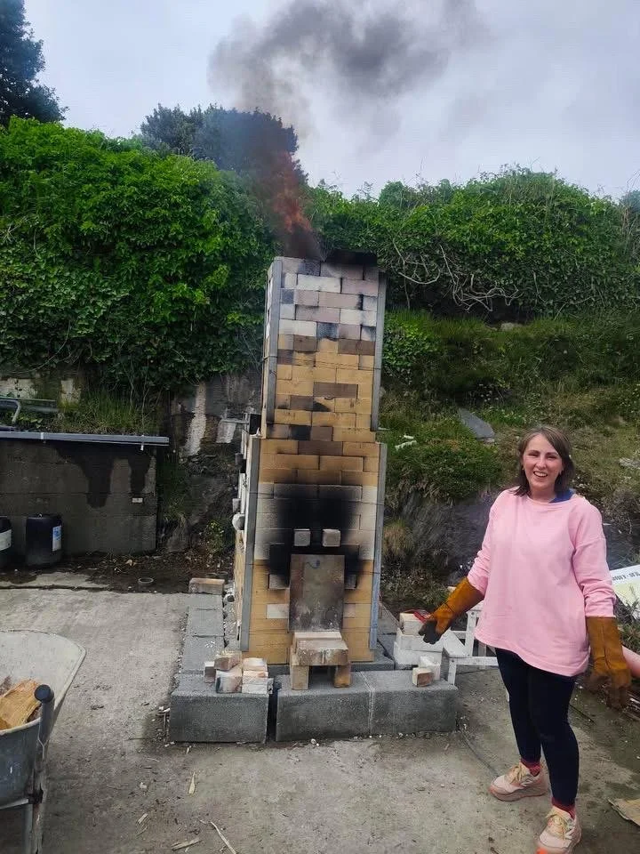 A woman standing next to a large outdoor brick oven on a concrete base, with smoke coming from the chimney, and her smiling and wearing gloves.