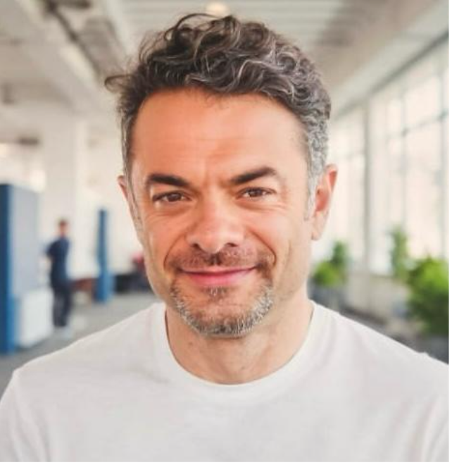A man with curly dark hair and a short beard smiling in a modern, well-lit indoor space with large windows and some plants in the background.