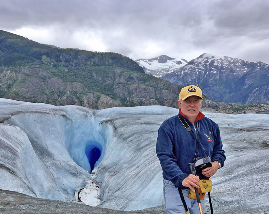 A man standing in front of a glacier with a mountain range in the background, wearing a yellow cap with 'Cal' written on it, a blue jacket, and holding a camera.