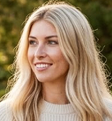 A smiling woman with long blonde hair outdoors, with greenery in the background.