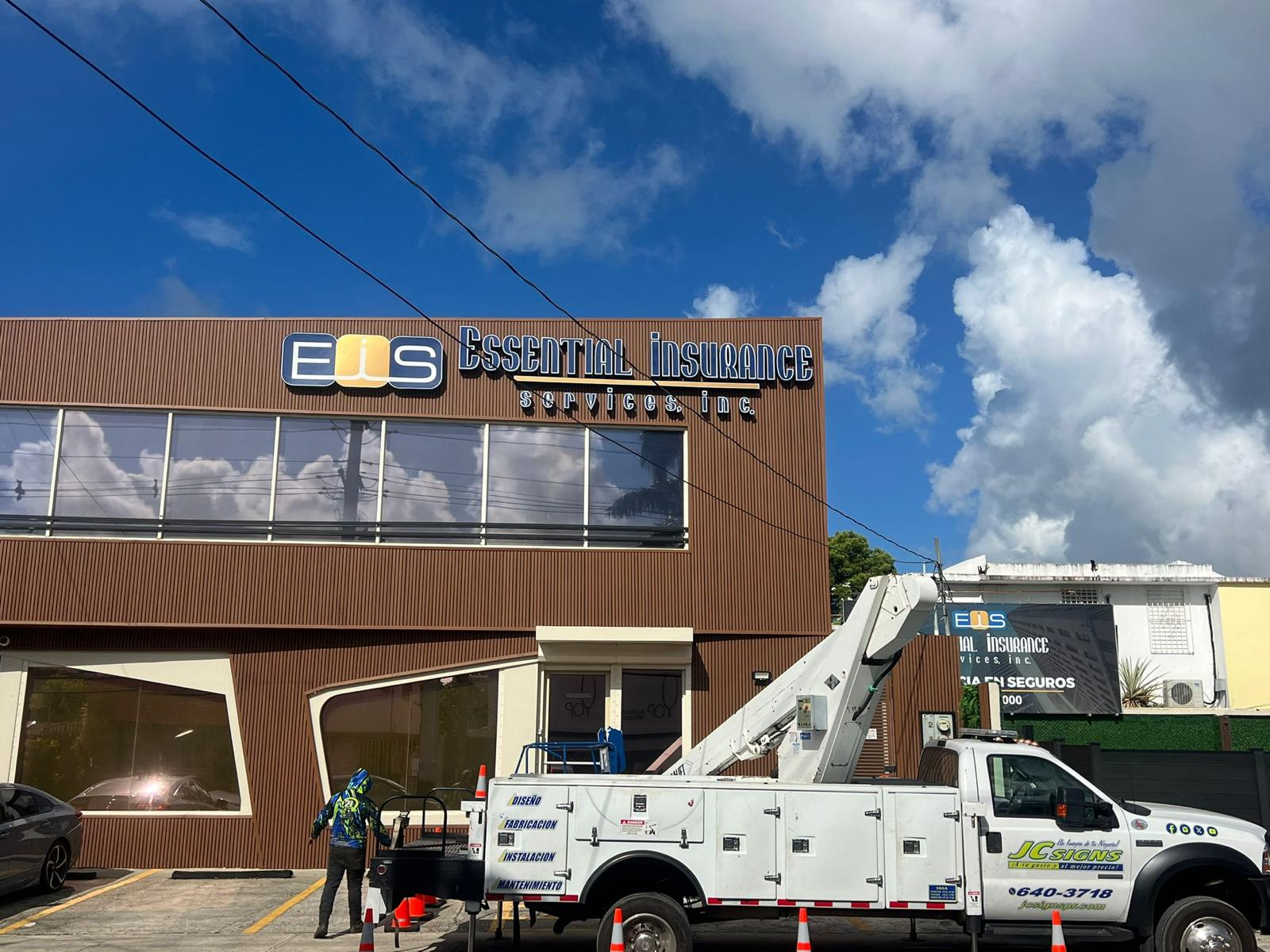 Edificio con letrero de 'Essential Insurance Services Inc.' y un camión de trabajo en el estacionamiento. Cielo despejado con nubes blancas y algunas líneas eléctricas.