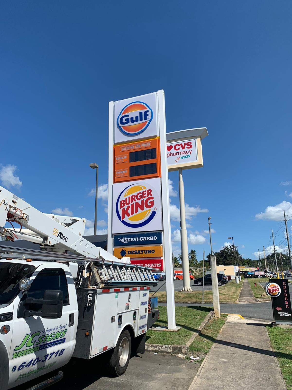 Un cartel de gasolina de Gulf, un restaurante Burger King, una farmacia CVS, y un camión de servicios con equipo en la calle, en un día soleado con cielo despejado.