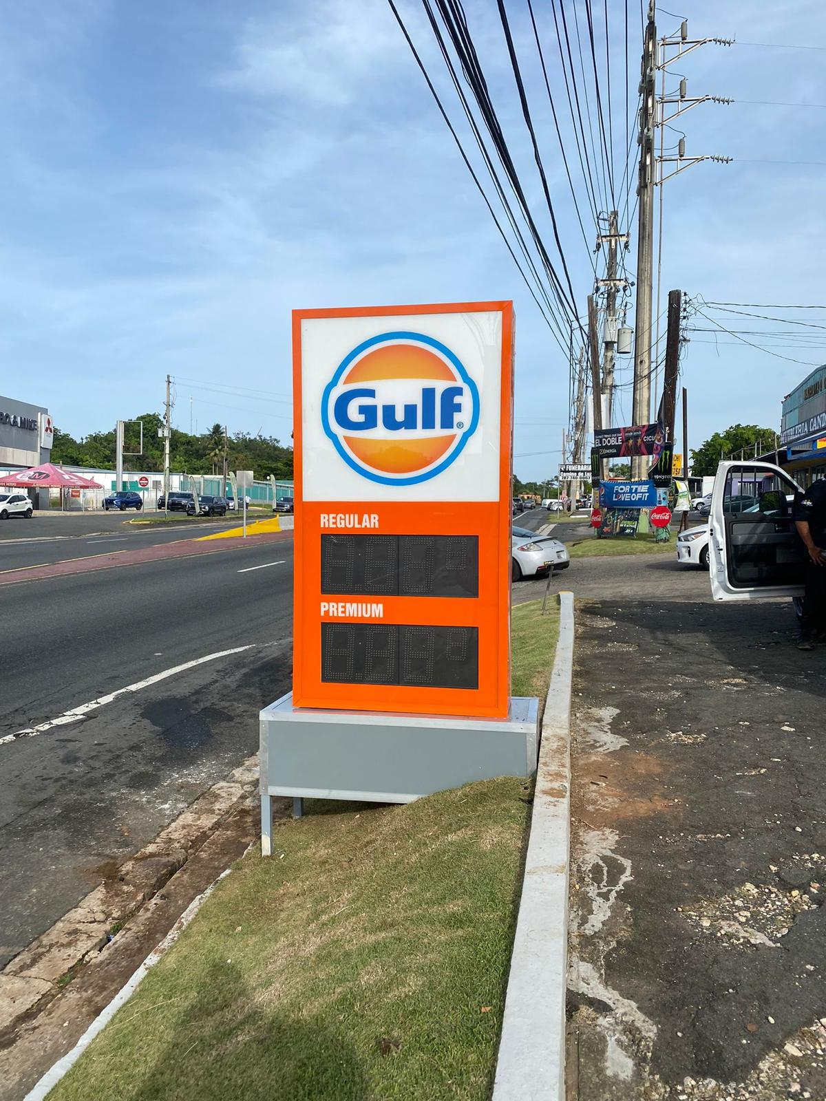 Puesto de gasolina Gulf con tarifas de gasolina regular y premium en una vía de tránsito en un día soleado.