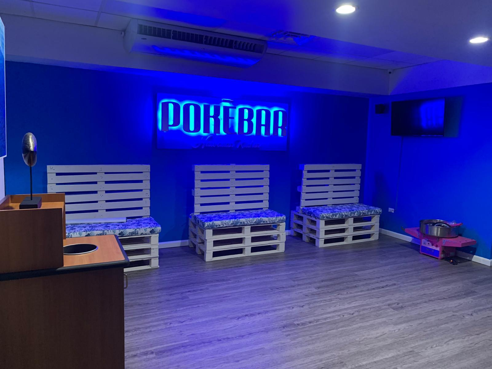 Sala con paredes azules, cartel luminoso que dice 'POKEBAR', tres bancos de madera pintados de blanco con almohadones, y una televisión en la esquina, ambiente de bar o sala de juegos.