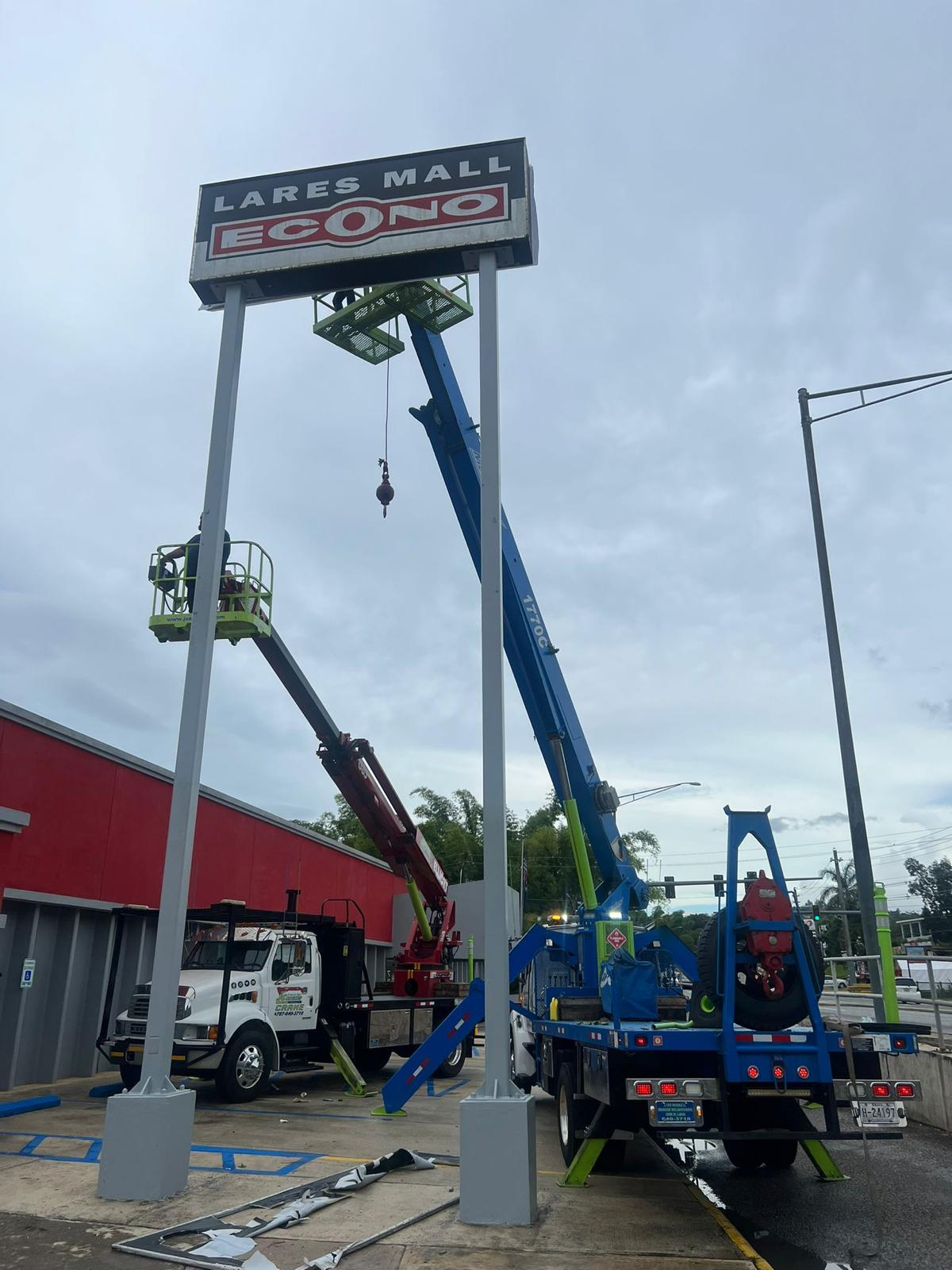 Trabajadores en plataformas elevadoras instalando una señal de letrero de LARES MALL y ECO NO en un poste alto, en un estacionamiento con herramientas y vehículos alrededor.