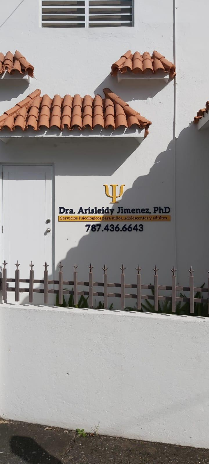 fachada blanca con un cartel de consulta psicológica, un símbolo psi, nombre de la doctora Arisleidy Jiménez, PhD, y teléfono, con un cielo soleado y sombra en la pared