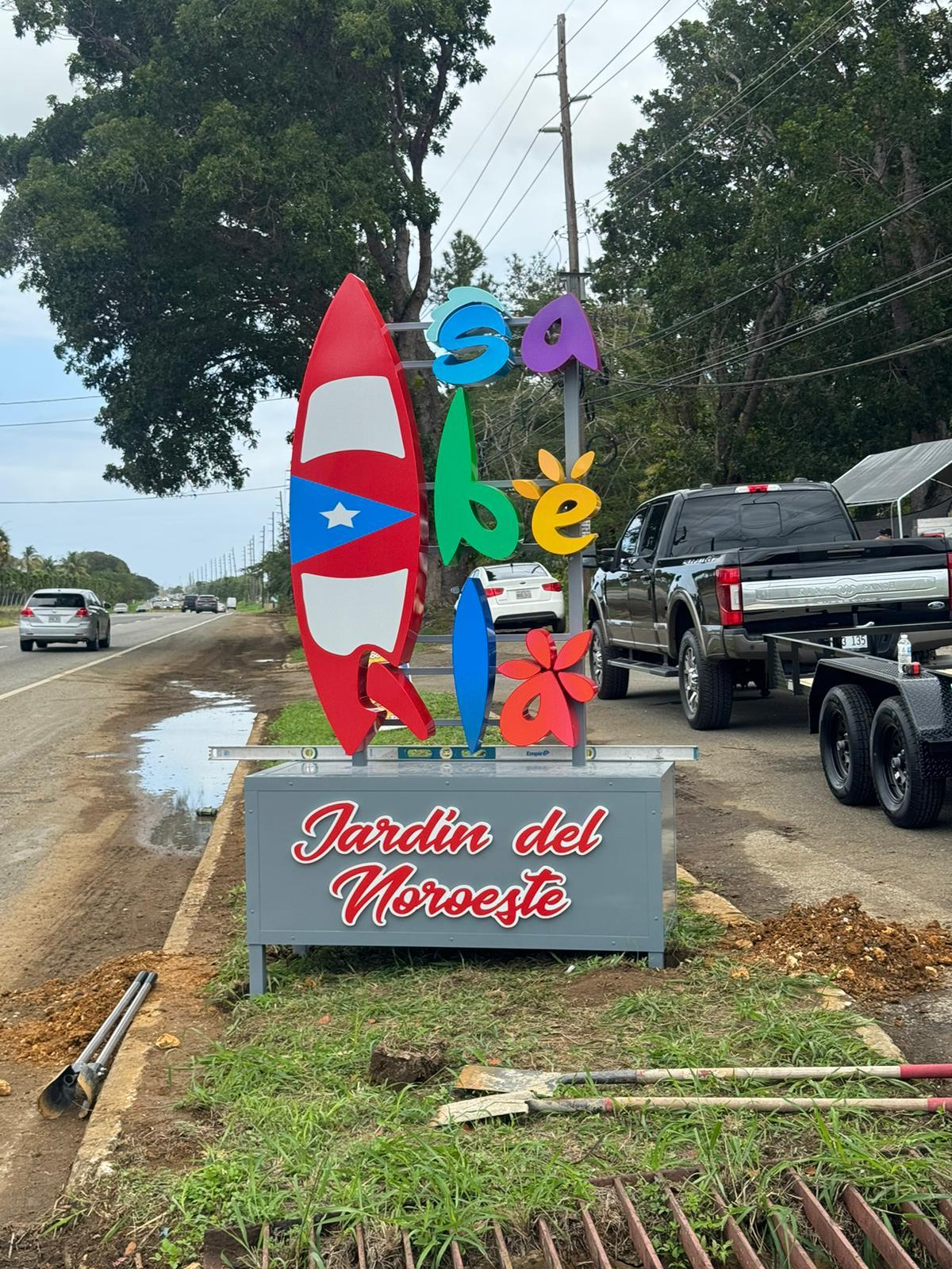 Cartel decorativo en la carretera con palabras en colores vivos que dicen 'Saba be', acompañado de una figura con los colores y símbolo de la bandera de Puerto Rico, con un texto en la base que dice 'Jardín del Noroeste'.