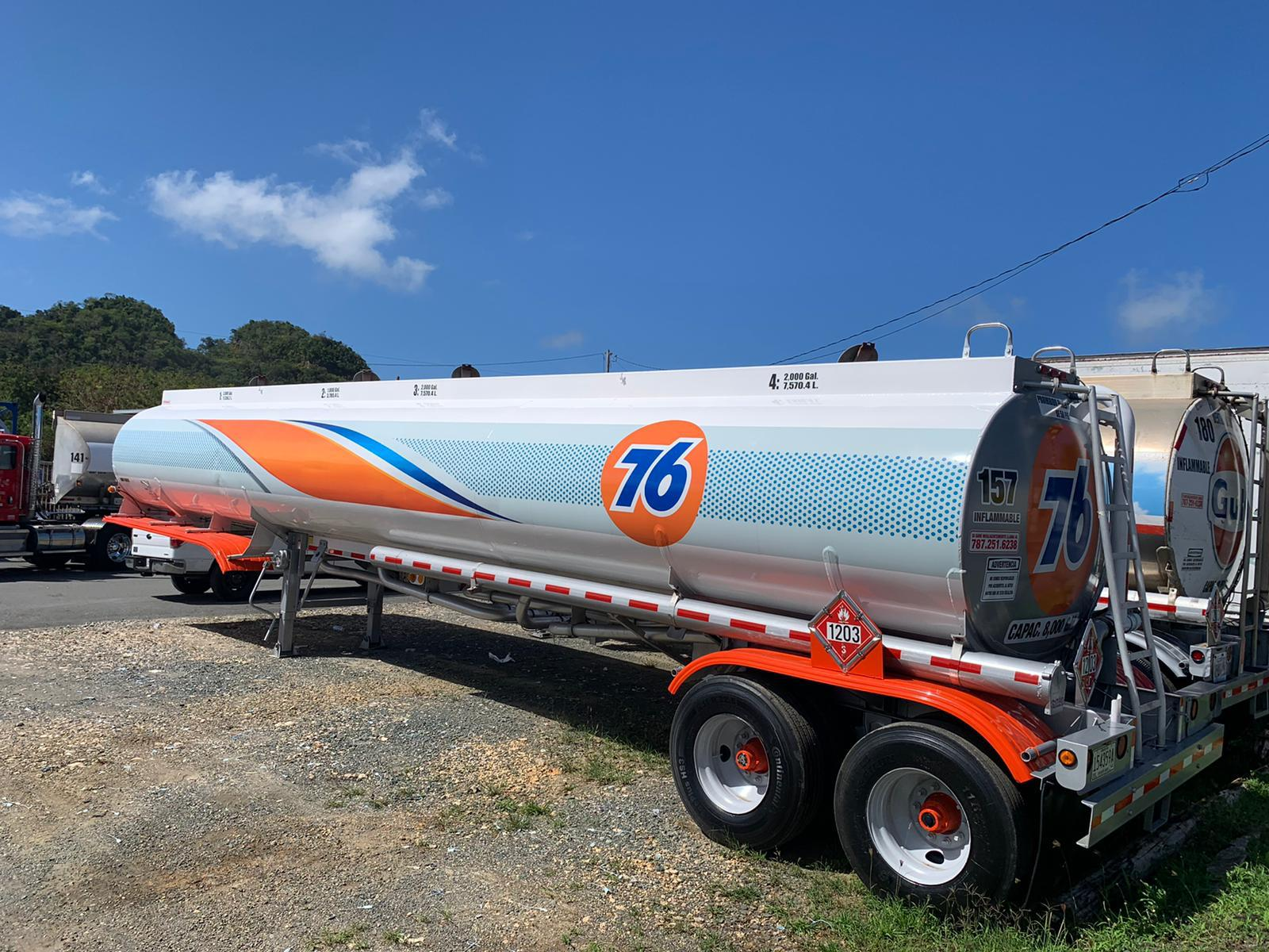 Tanque de gasolina con logotipo de marca 76, sobre remolque en área de estacionamiento, con otros vehículos y montañas al fondo, cielo despejado.