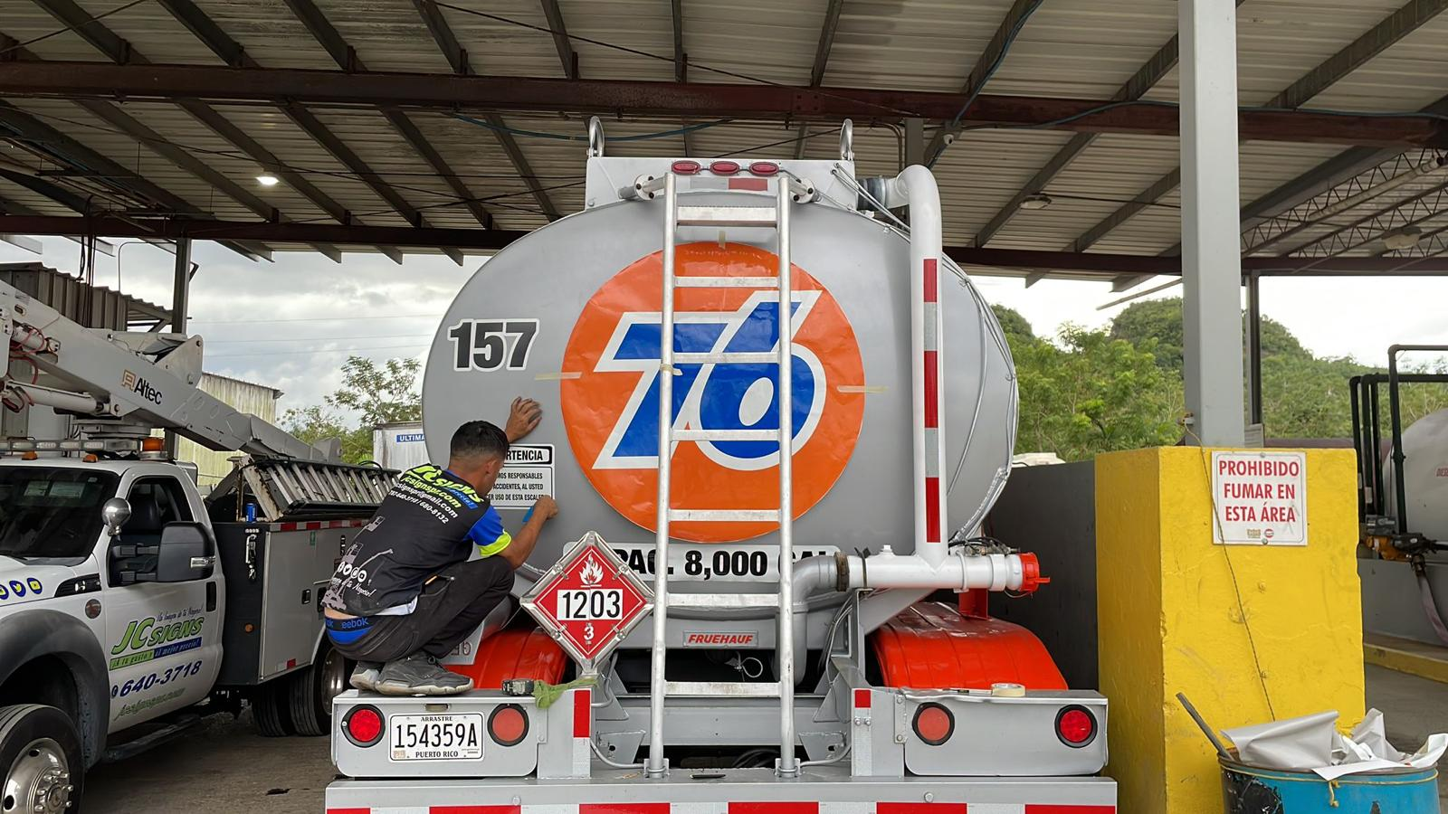 Un camión cisterna de gasolina en un área cubierta, con un técnico escribiendo en la parte trasera. Hay una señal que prohíbe fumar en la zona y un vehículo de servicios junto a la cisterna.
