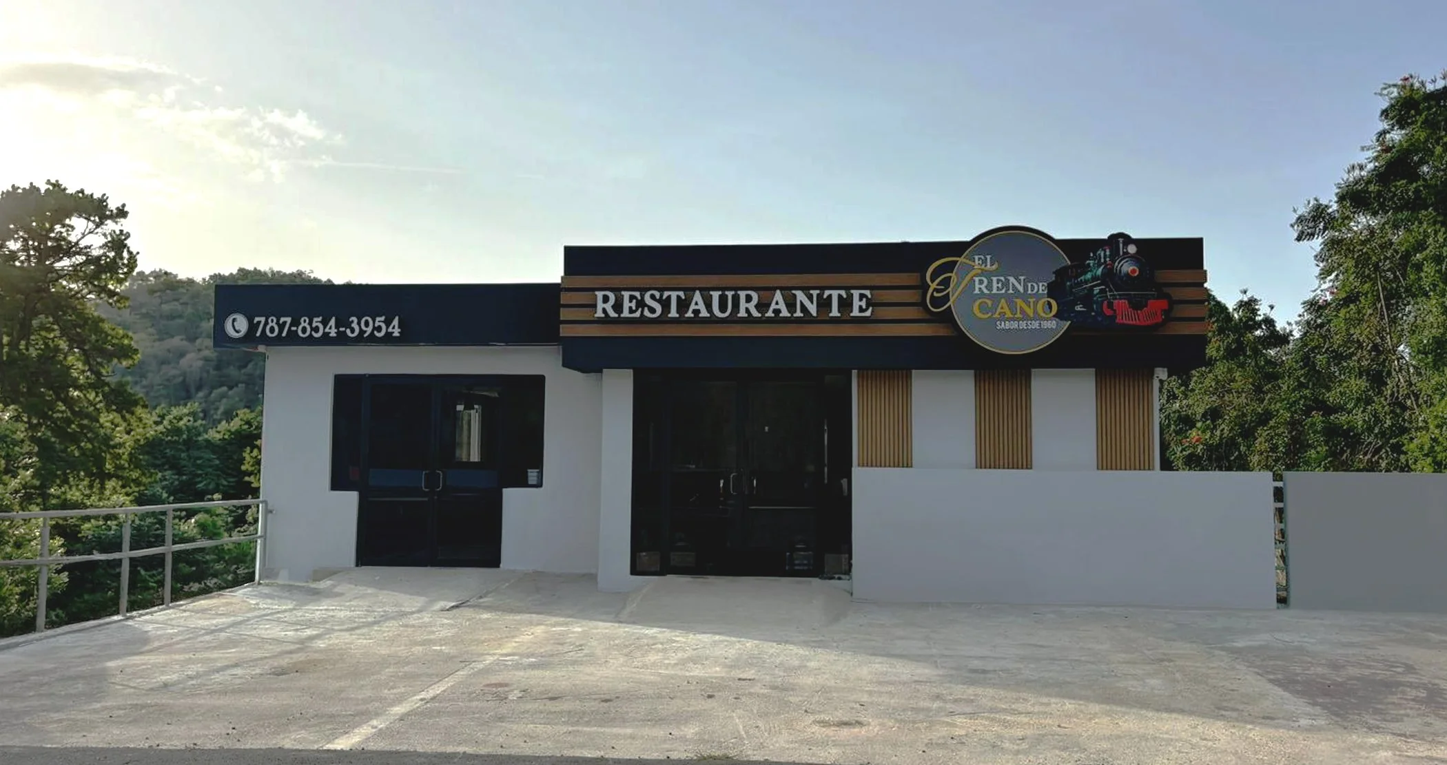 Fachada de un restaurante llamado 'El Tren de Cano', con un dibujo de un tren y un número de contacto 787-854-3954, rodeado de árboles verdes y un cielo despejado.