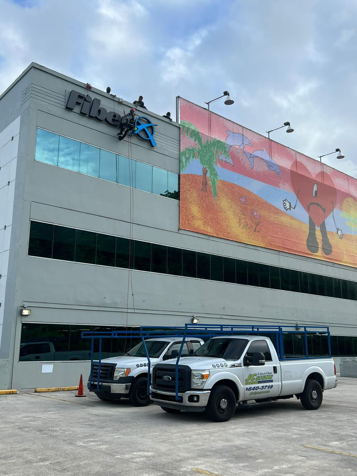 Trabajo de mantenimiento en la fachada de un edificio, con un trabajador en andamios y dos personas en el techo, junto a un mural colorido con una palmera, delfines y una figura con corazón y zapatos.