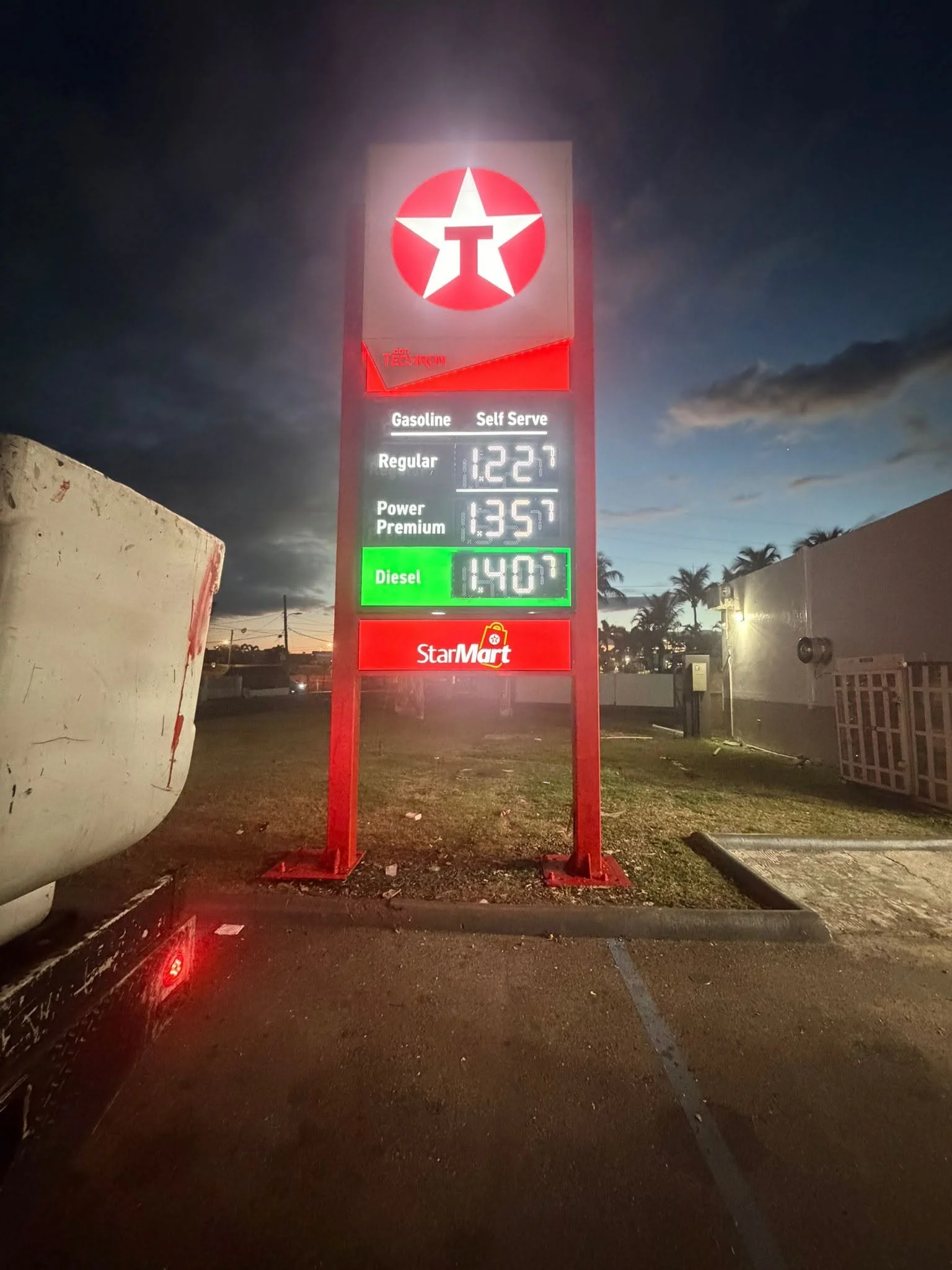 Pantalla de gasolina en un expendio de combustible en la noche, mostrando precios para gasolina regular, Power Premium y diésel, en un fondo con cielo oscuro y algunas palmeras.