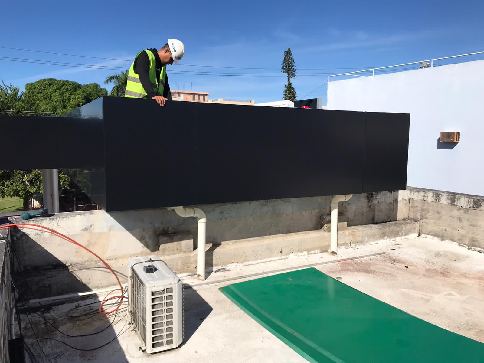 Un trabajador con casco y chaleco de seguridad inspecciona una estructura moderna negra en un edificio en construcción. En el suelo hay un aire acondicionado portátil, cables y una alfombra verde.