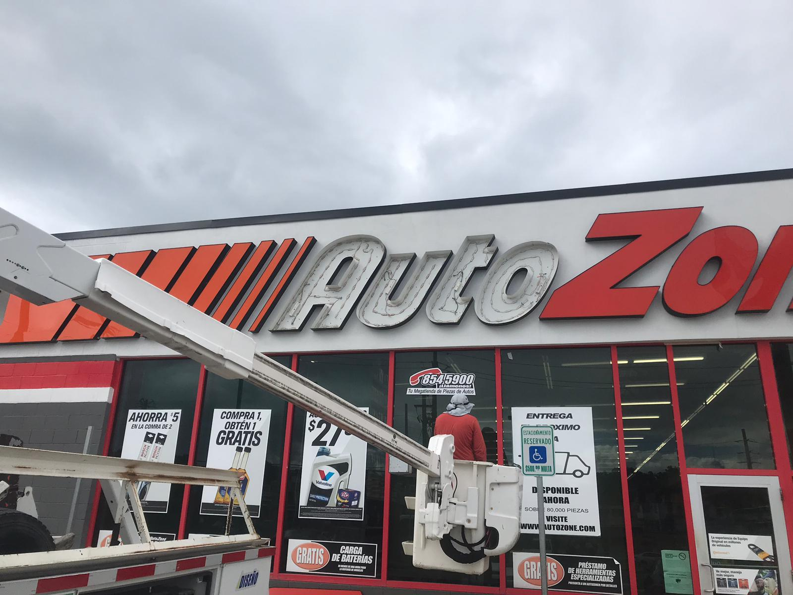 Letrero de la tienda AutoZone en un día nublado, vista desde el exterior, con vigas de elevación y empleados en la entrada.