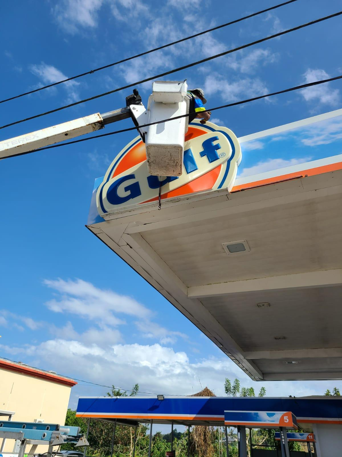 Trabajador en elevador limpiando un letrero de gasolina en una estación de servicio bajo un cielo azul con nubes.