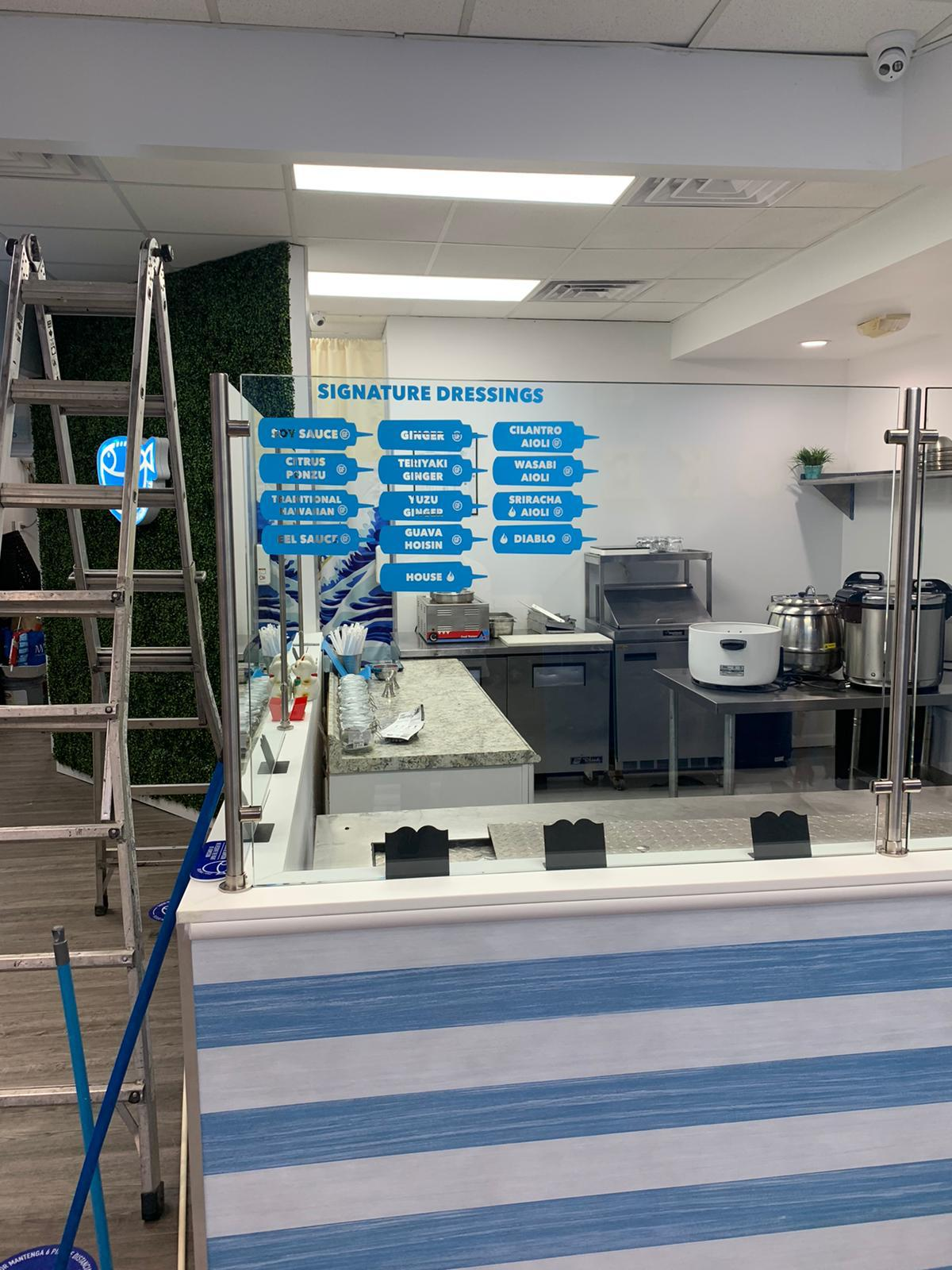Mostrador de comida con un cartel azul que lista salsas y aderezos con nombres en inglés y algunos en japonés, y una cocina en el fondo. Hay una escalera de aluminio a la izquierda y una pared decorativa con plantas verdes a la izquierda.