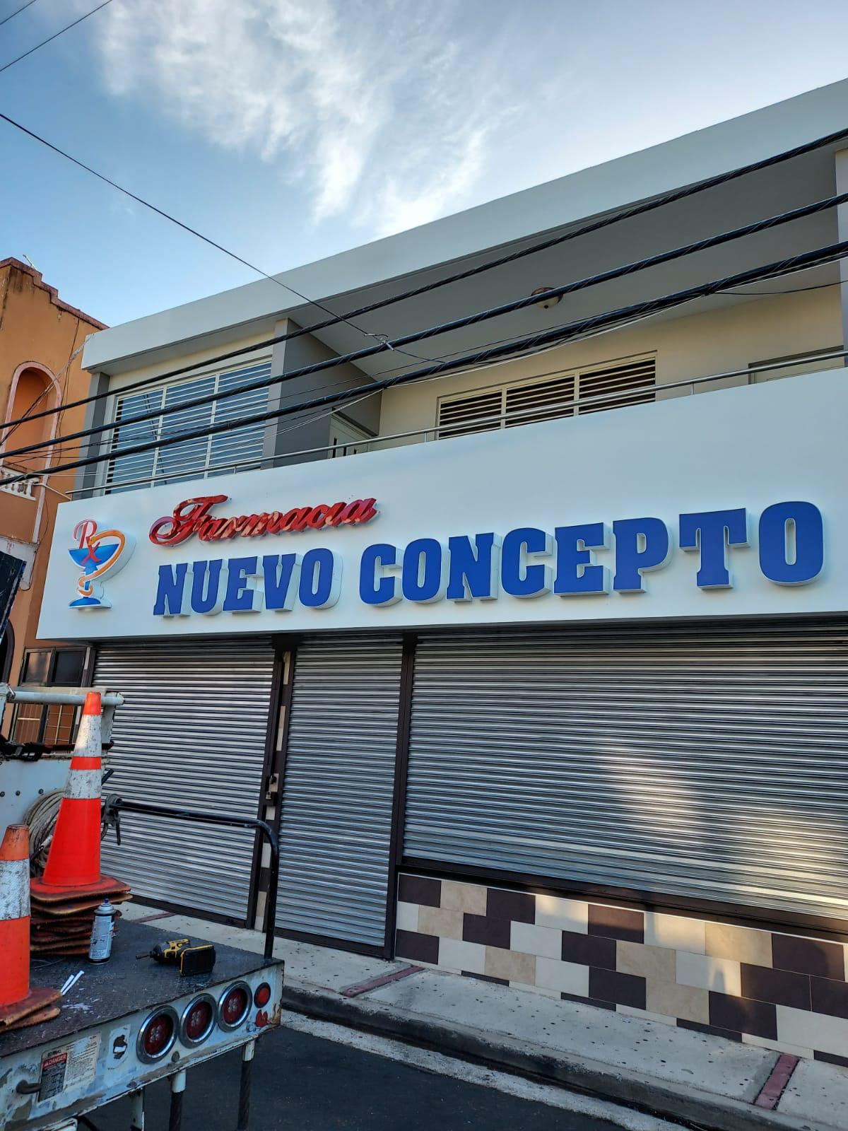 Fachada de una farmacia llamada 'Nuevo Concepto' con letras grandes en azul y un cartel en la parte superior que dice 'Farmacia' en letras rojas. Hay un logotipo con una copa y una letra 'R' en colores rojo y azul. La tienda tiene persianas metálicas