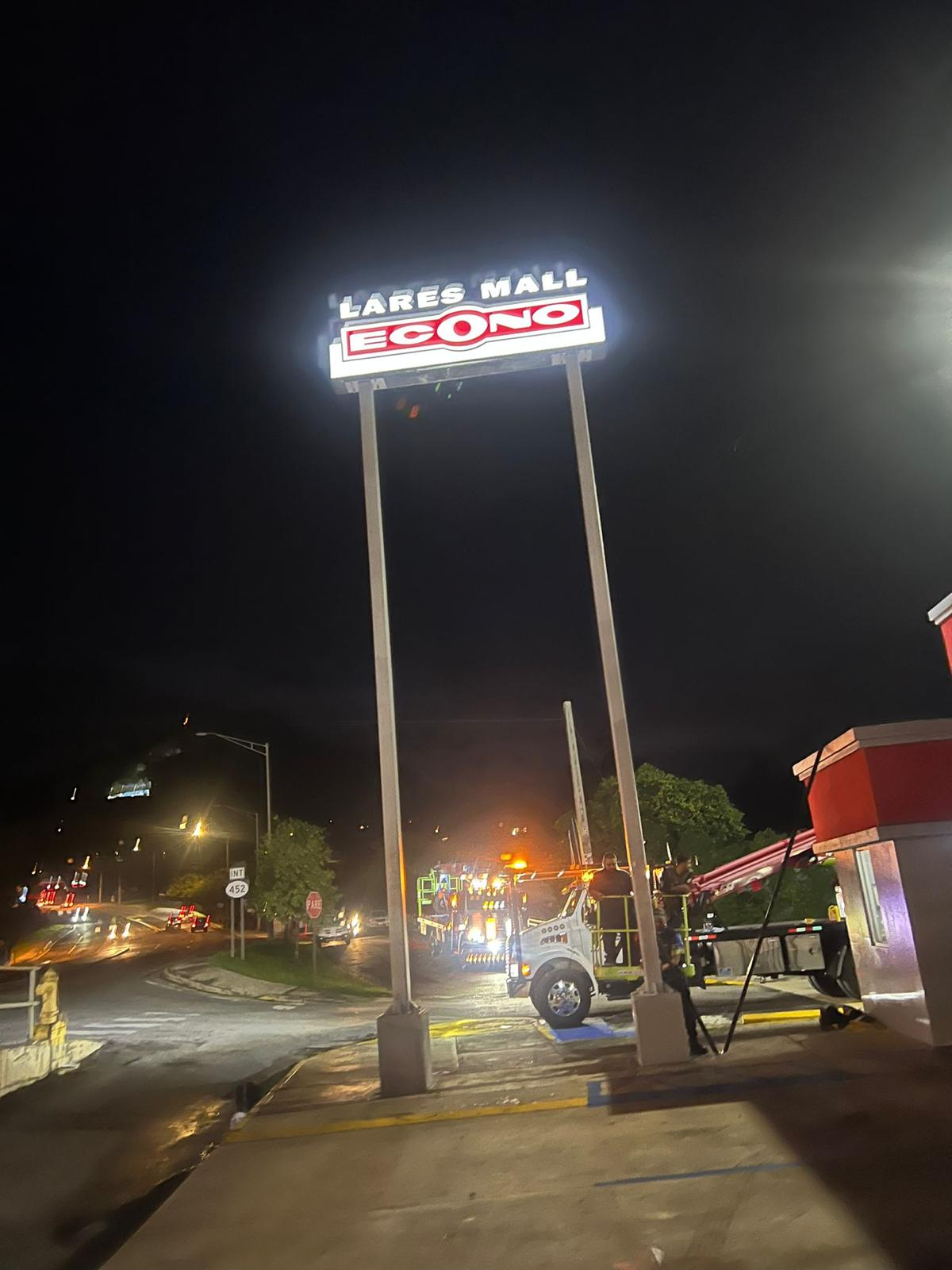 Vista nocturna de un letrero iluminado que dice 'Lares Mall Econo' y algunos vehículos de servicio y personas trabajando en la calle.