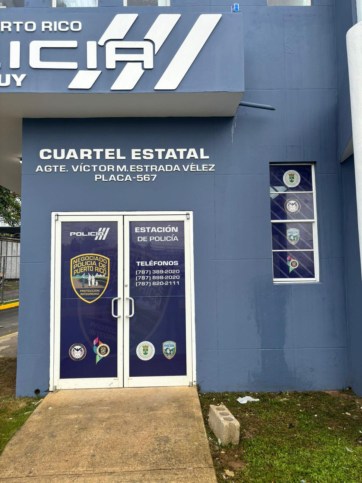 Edificio azul con señalización de una estación de policía en Puerto Rico, con el nombre de cuartel estatal, teléfonos de contacto y varios logotipos de diferentes departamentos policiales.