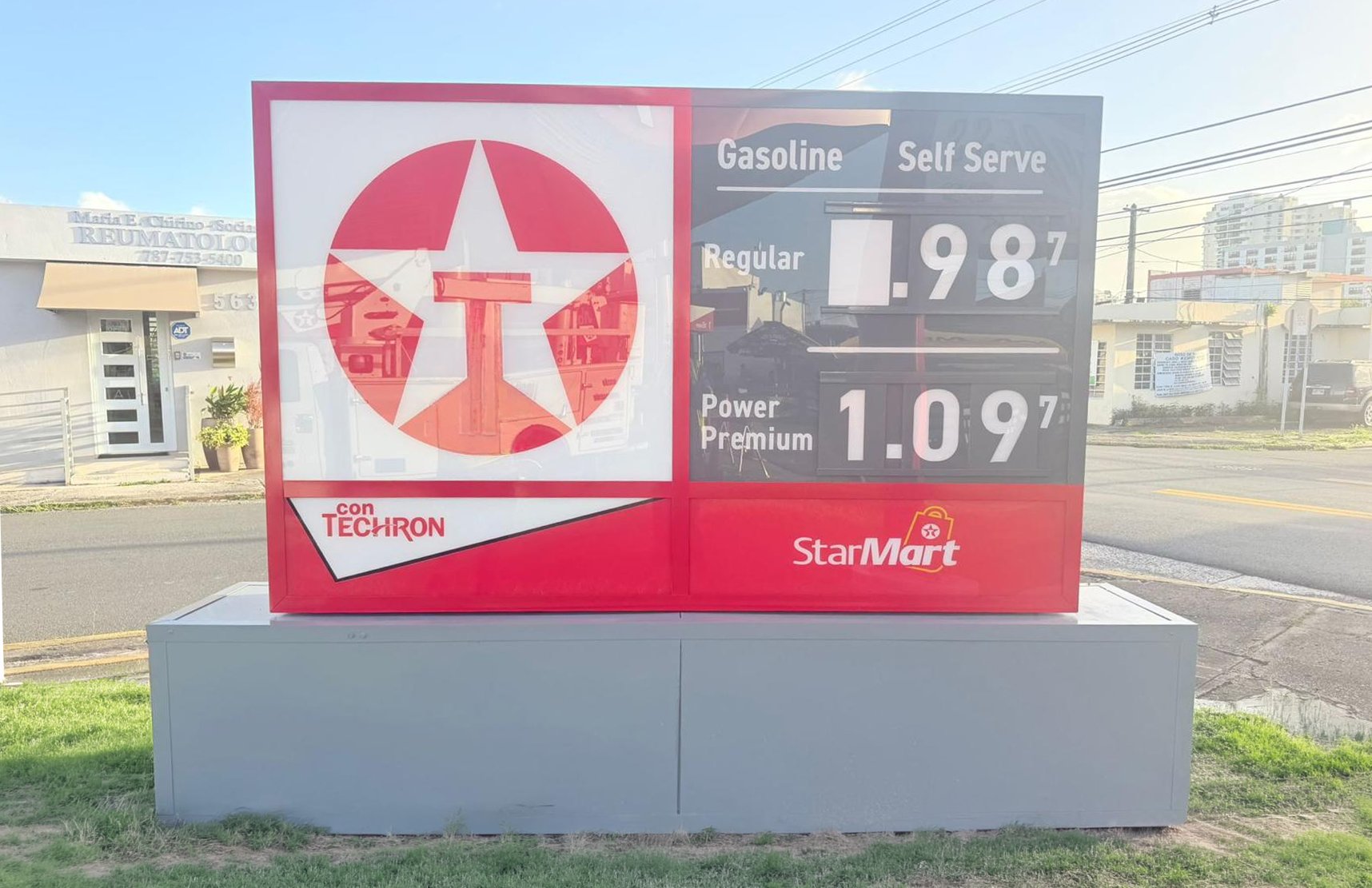 Cartel de gasolina en una estación de servicio que muestra los precios del combustible con marca StarMart, incluyendo gasolina regular a 1.98 y Power Premium a 1.09, con publicidad de Techron, en un entorno urbano con edificios y líneas eléctricas.
