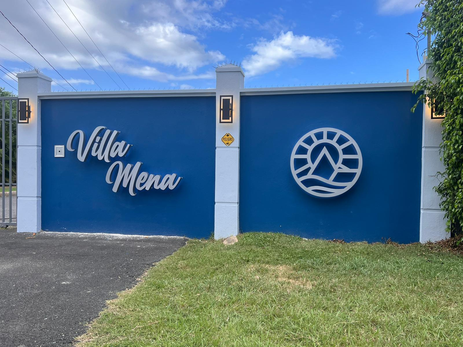 Muro azul con letras blancas que dicen 'Villa Mena' y un logotipo en forma de círculo con un árbol y olas en su interior, en una entrada de propiedad.