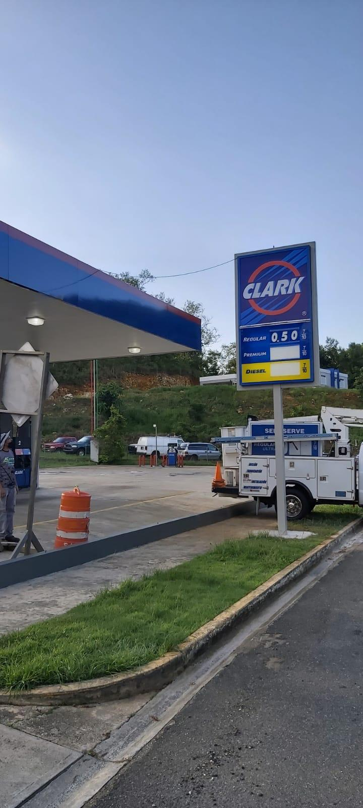 Vista de una gasolinera Clark con un cartel que muestra los precios de combustible, con una área de estacionamiento y un área de servicio, algunos autos estacionados y un cielo despejado.
