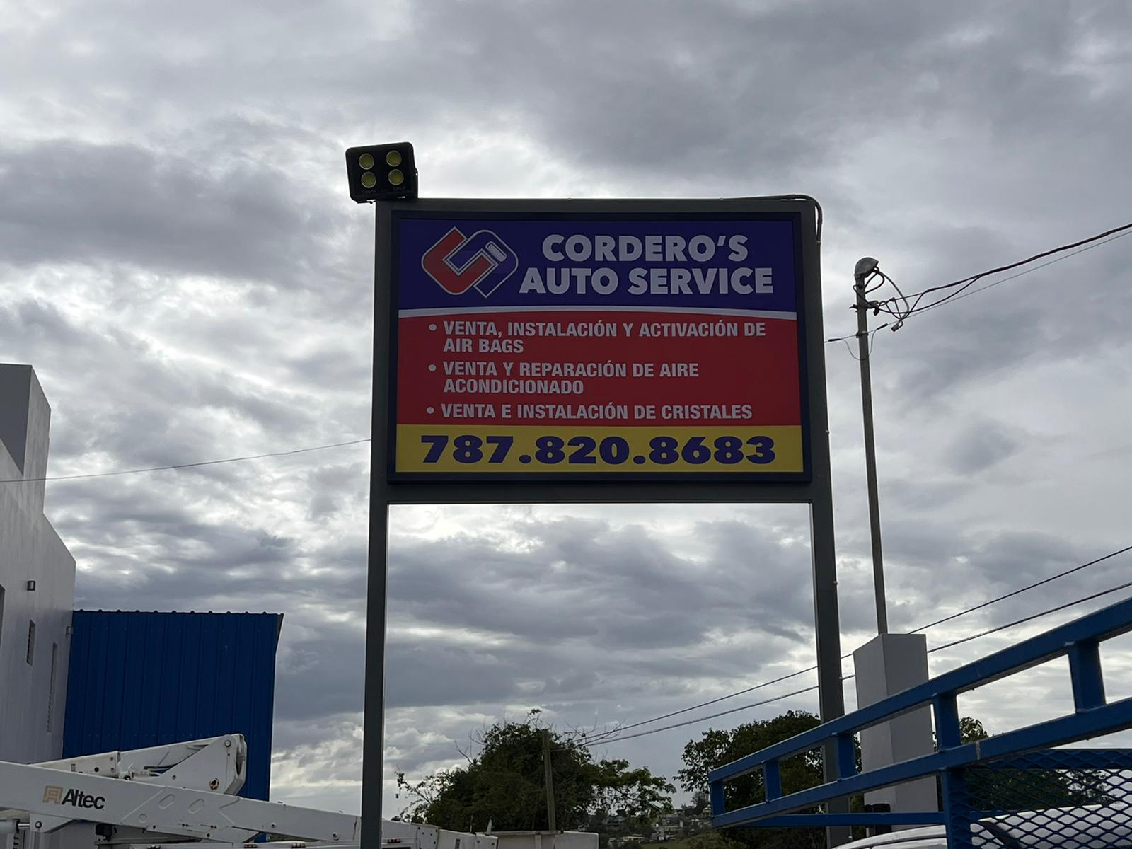 Cartel publicitario de Cordero's Auto Service con información de servicios y número de contacto, contra fondo de cielo nublado y cables de electricidad.