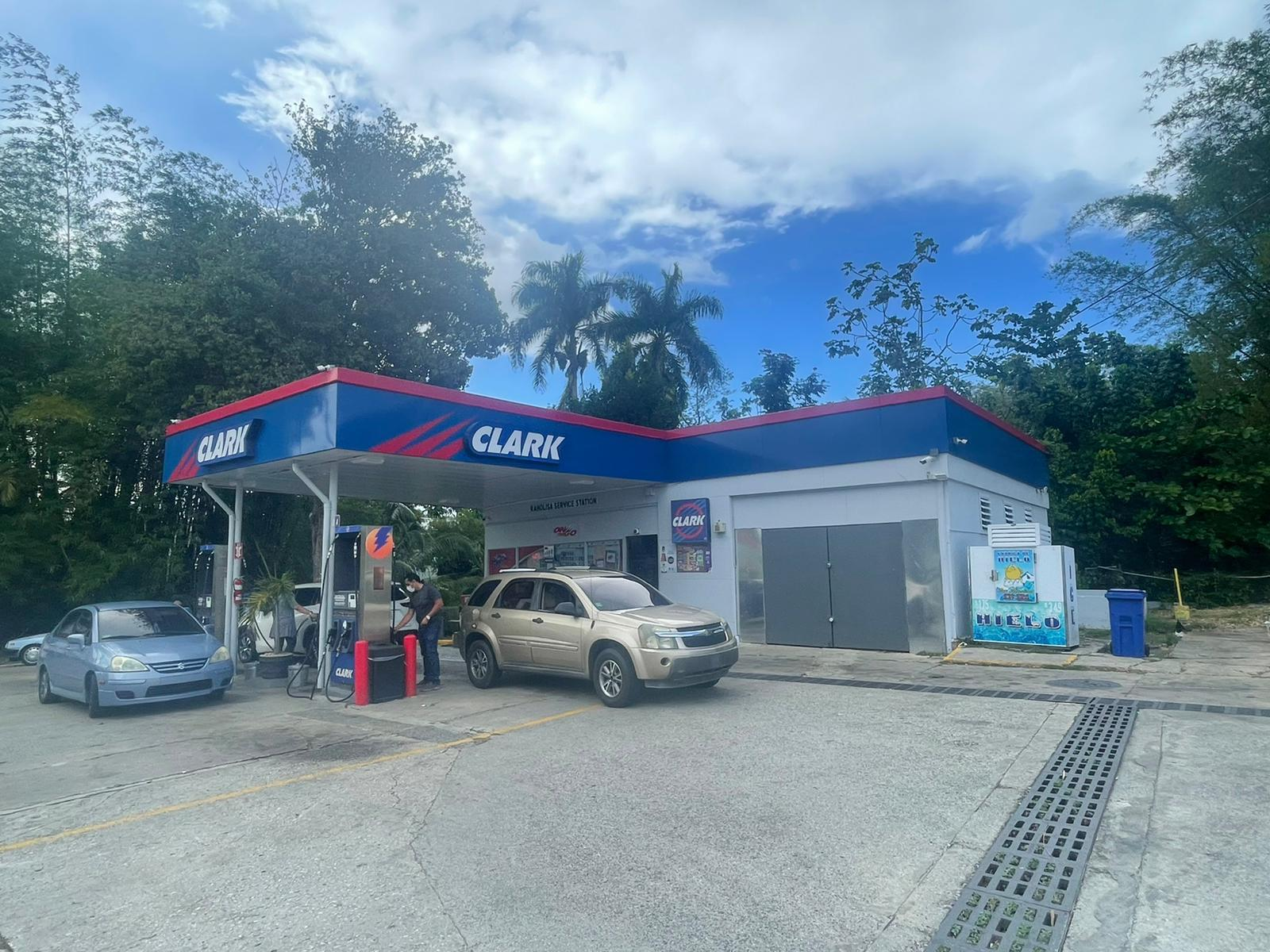 Estación de servicio de gasolina con una gasolinera Clark, dos autos en la estación y dos personas abasteciendo gasolina. Al fondo hay árboles y cielo con algunas nubes.
