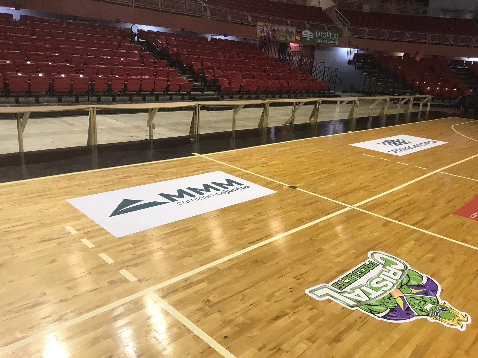 Interiores de un gimnasio con un piso de madera y asientos rojos en las gradas. En el suelo, en primer plano, hay un logotipo de los New York Knicks, otro de los Dreamers y anuncios publicitarios en la cancha.
