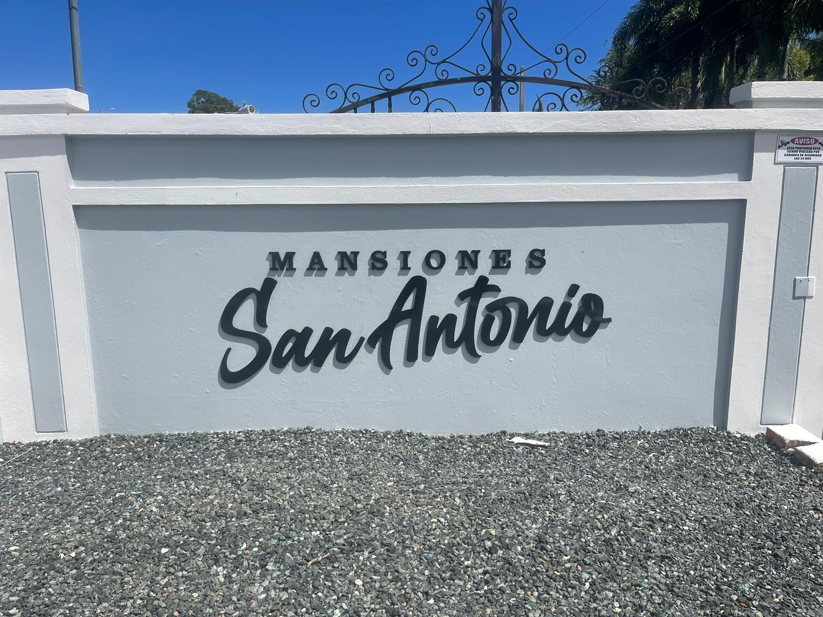 Muro con cartel que dice 'Mansiones San Antonio' en letras negras y grandes, en un fondo de color blanco y con piso de grava en primer plano.