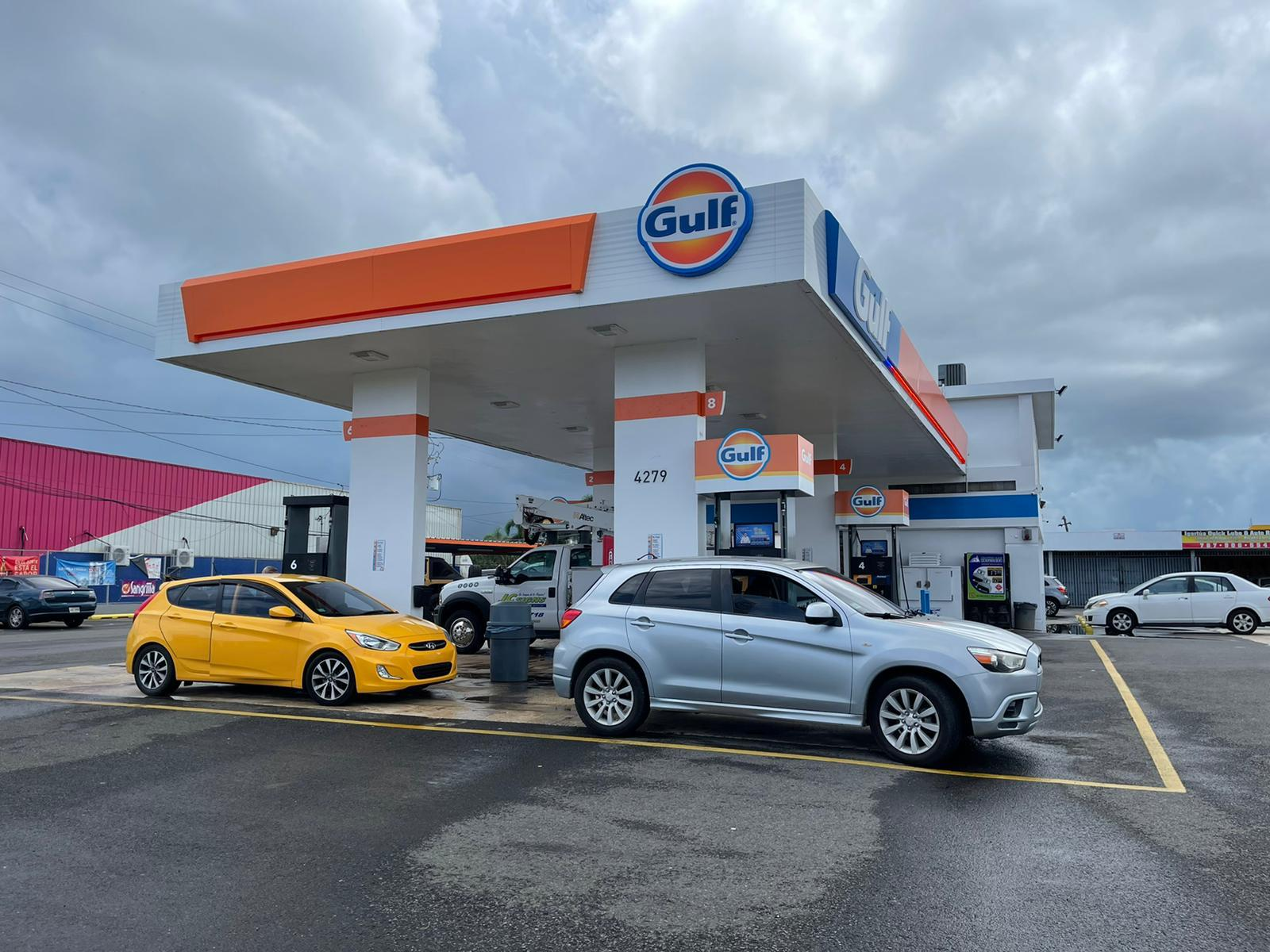 Estación de servicio de gasolina Gulf con varias personas y autos esperando, cielo nublado.