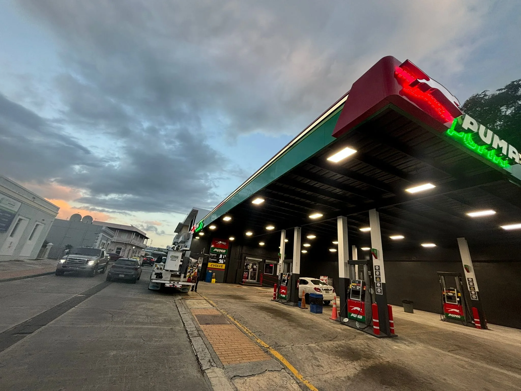 Una estación de servicio con bombas de gasolina y vehículos estacionados, bajo un cielo nublado en horas de la tarde o atardecer.