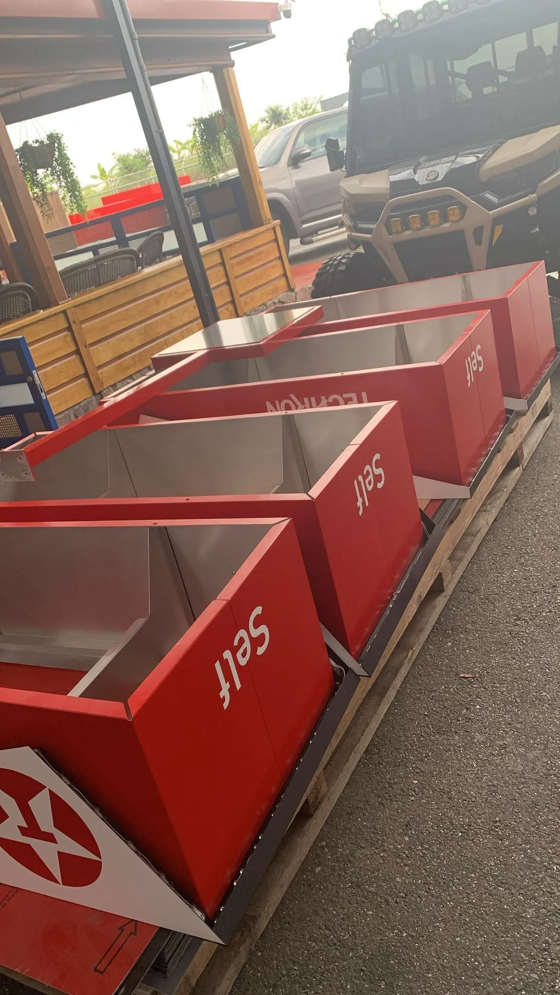 Contenedores rojos en una estructura de madera, con la palabra 'self' en ellos, en un lugar abierto con vehículos estacionados al fondo.