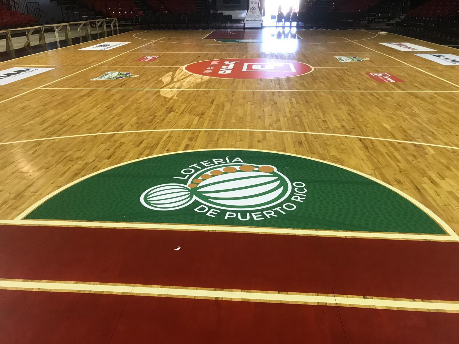 Vista de una cancha de baloncesto con el logotipo de la Liga de Puerto Rico en primer plano y anuncios publicitarios en el piso, con un grupo de personas al fondo en la entrada del lugar.