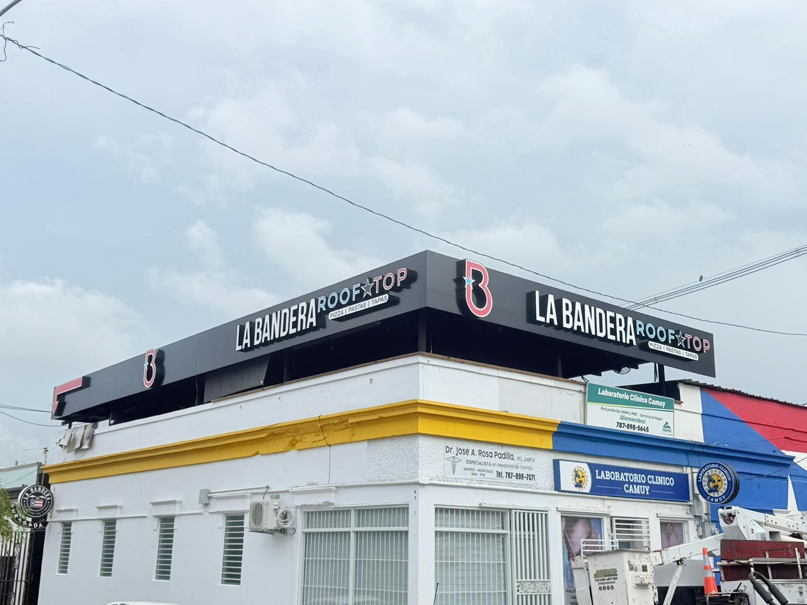 Fachada de un edificio con señalización de 'La Bandera Roof Top' que ofrece pizza, pastas y tapas, y un cartel de un laboratorio clínico llamado 'Laboratorio Clínico Camuy'.