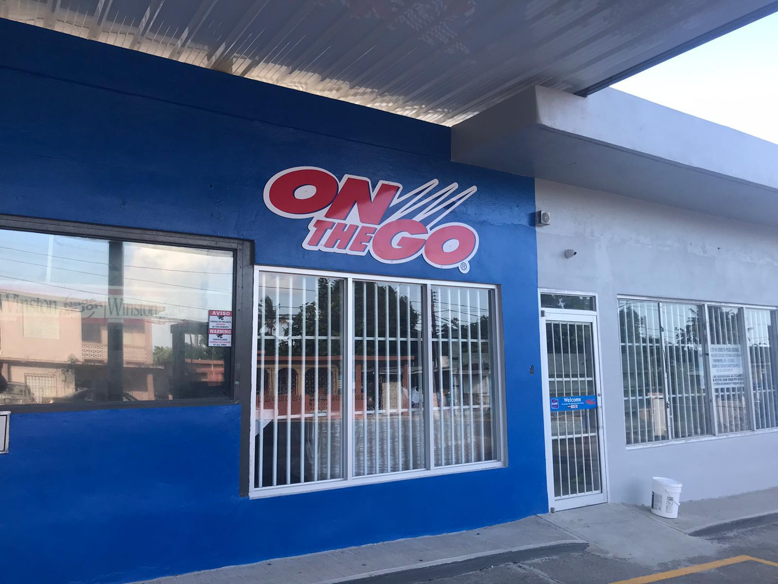 Fachada de un establecimiento con letrero que dice 'ON THE GO' en letras grandes rojas y blancas, con estructura de metal y ventanas con rejas.