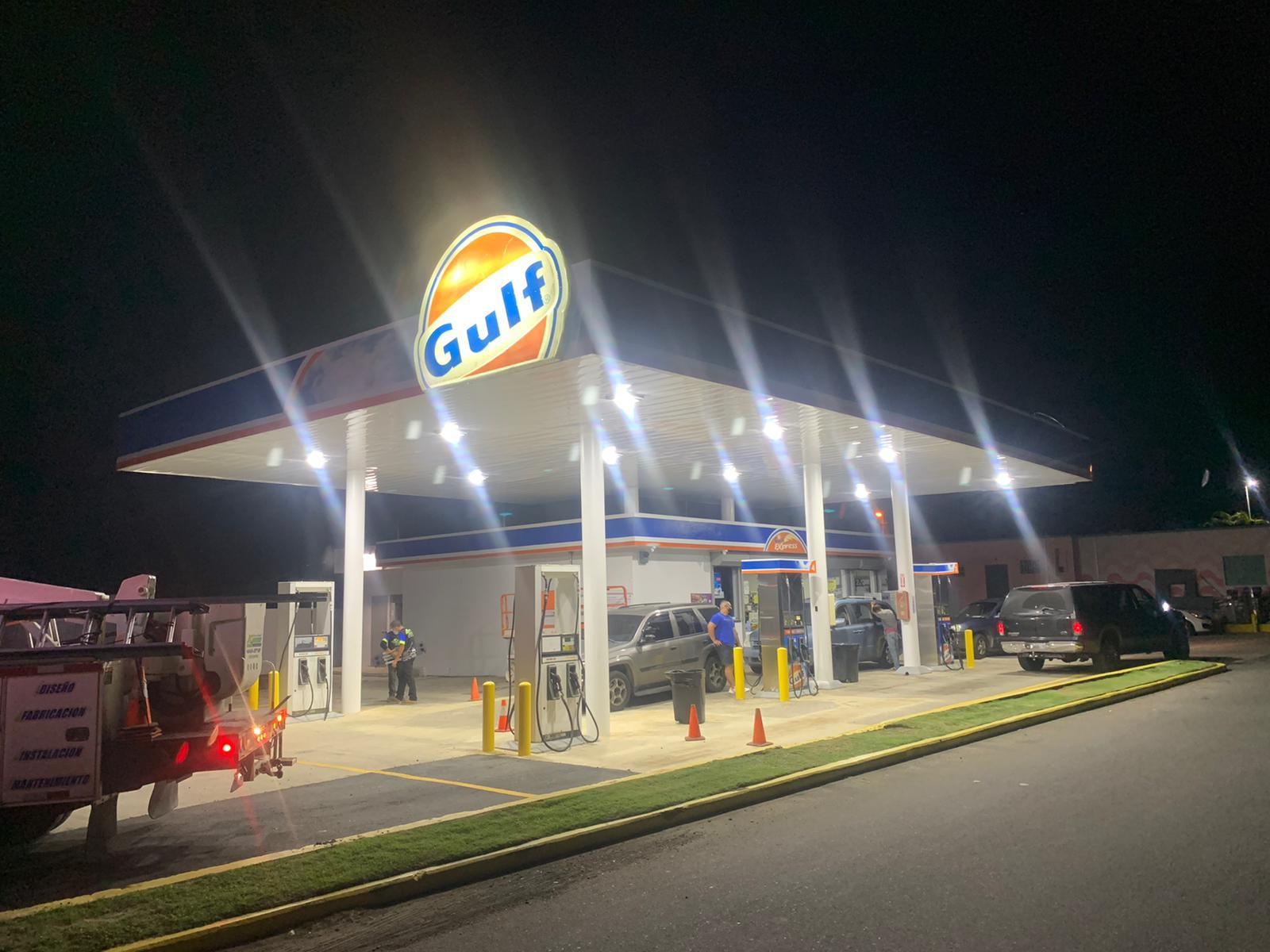 Estación de gasolina con logo de Gulf en la noche, con varias personas y vehículos. Iluminación brillante en el área de servicios.