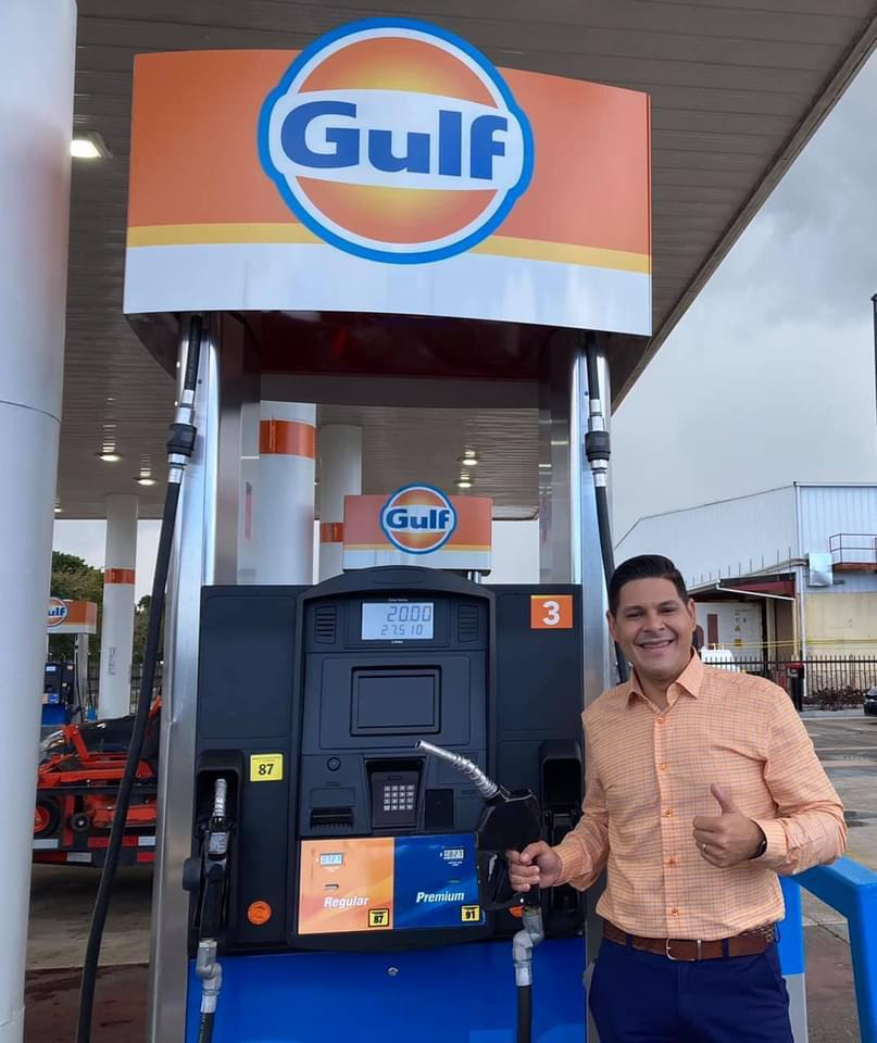 Imagen de una gasolinera Gulf con un hombre sonriendo y haciendo un gesto de aprobación, sosteniendo la boquilla de la bomba de gasolina.