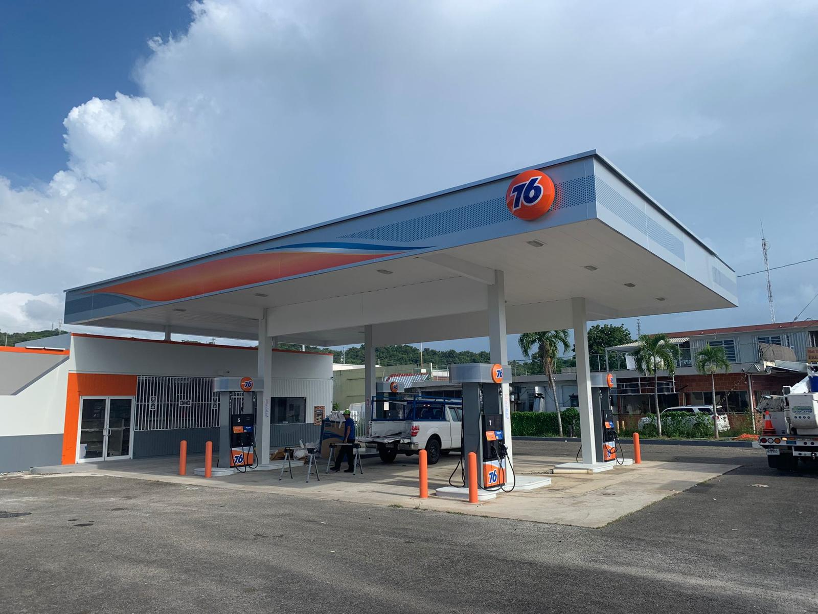 Estación de servicio de gasolina con banderas y bombas de combustible de la marca 76, ambiente de día soleado y edificio con árboles en el fondo.