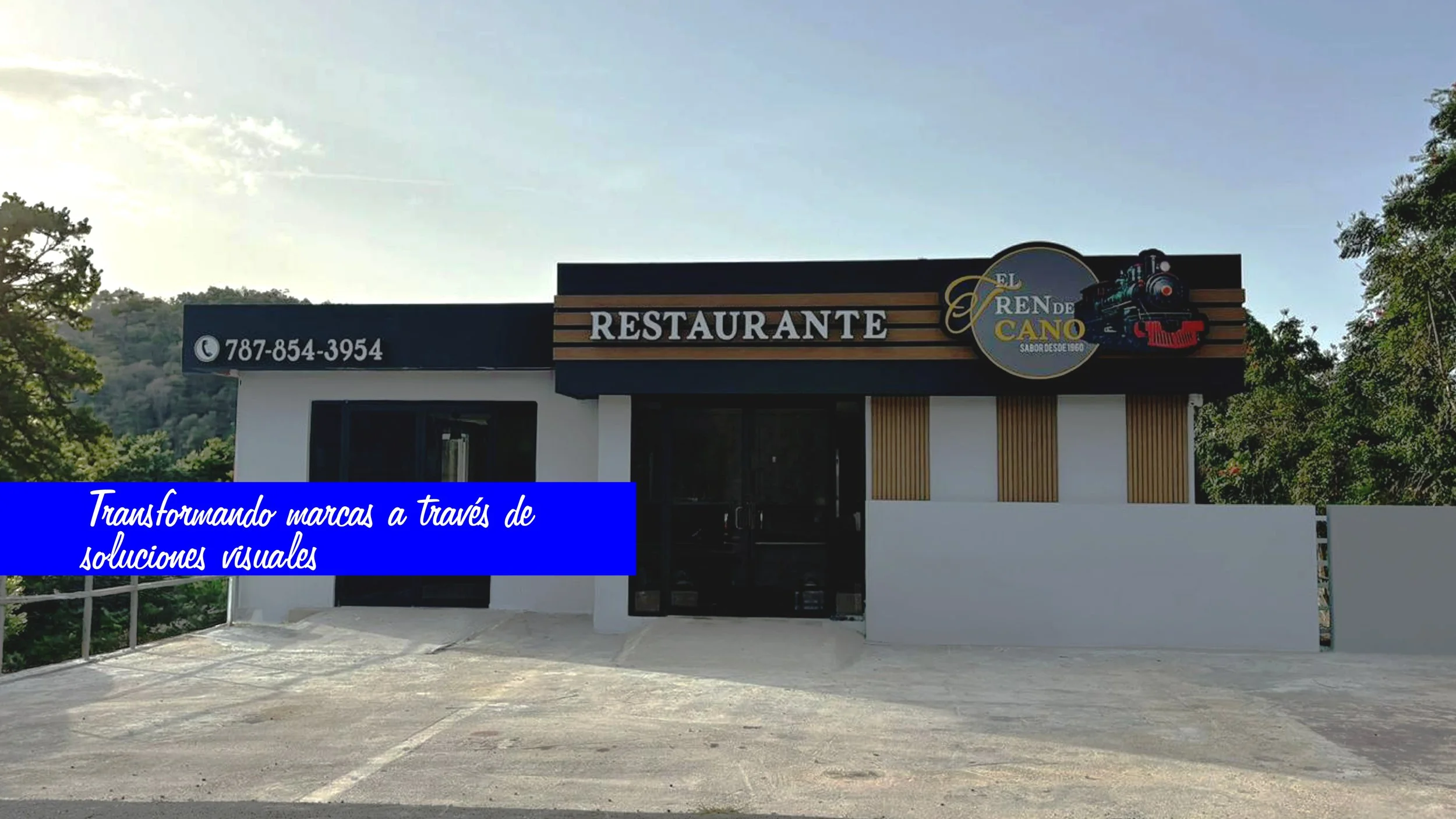 Fachada de un restaurante llamado El Tren de Cano, con un logotipo que presenta una locomotora de tren en colores negro, rojo y azul. El número de contacto en la pizarra, con fondo azul, 787-854-3954, y un mural decorativo con madera y paredes blancas.