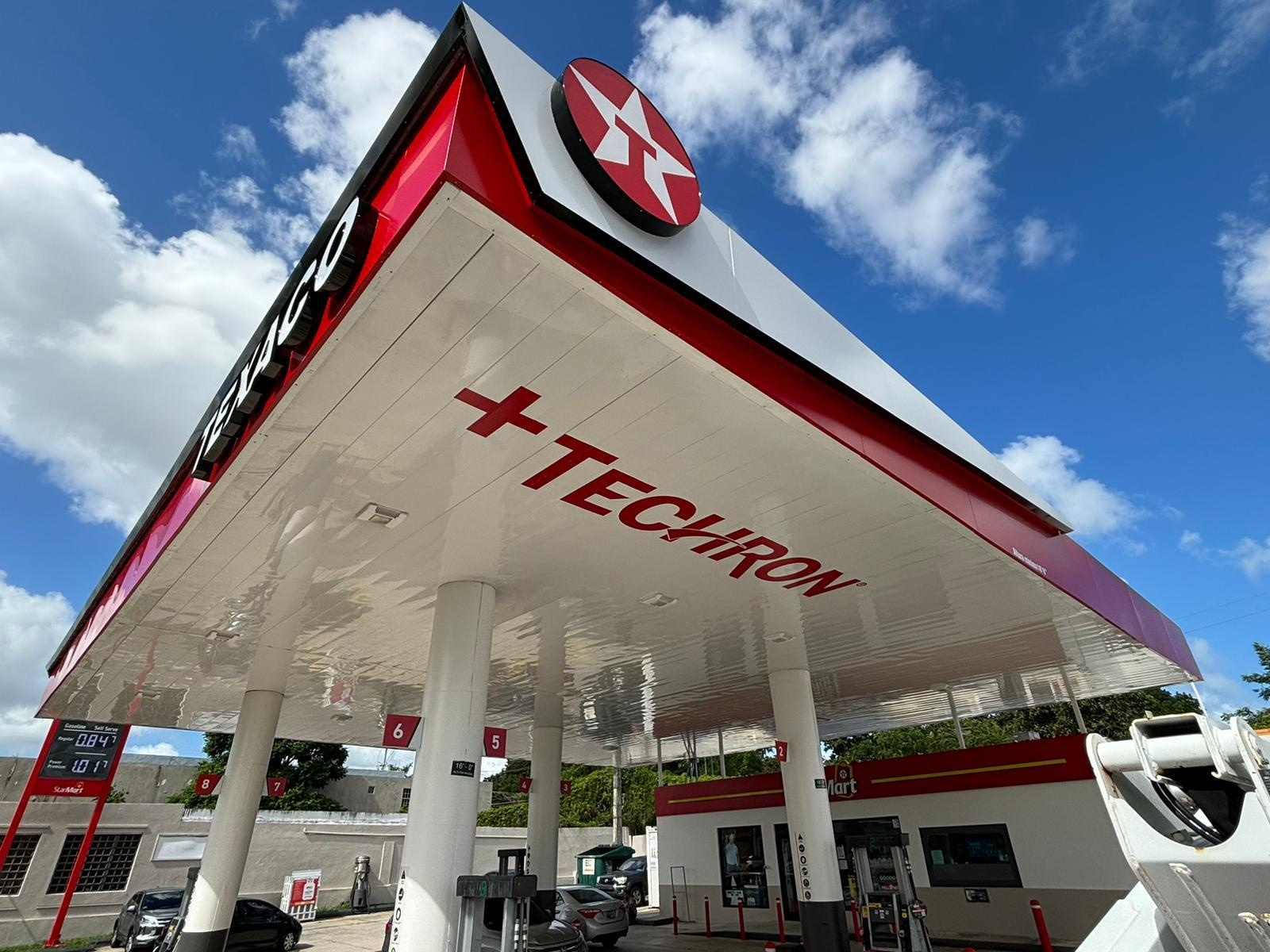 estación de gasolina Texaco con estructura moderna y bandera en la parte superior, bajo un cielo azul con nubes, en un día soleado